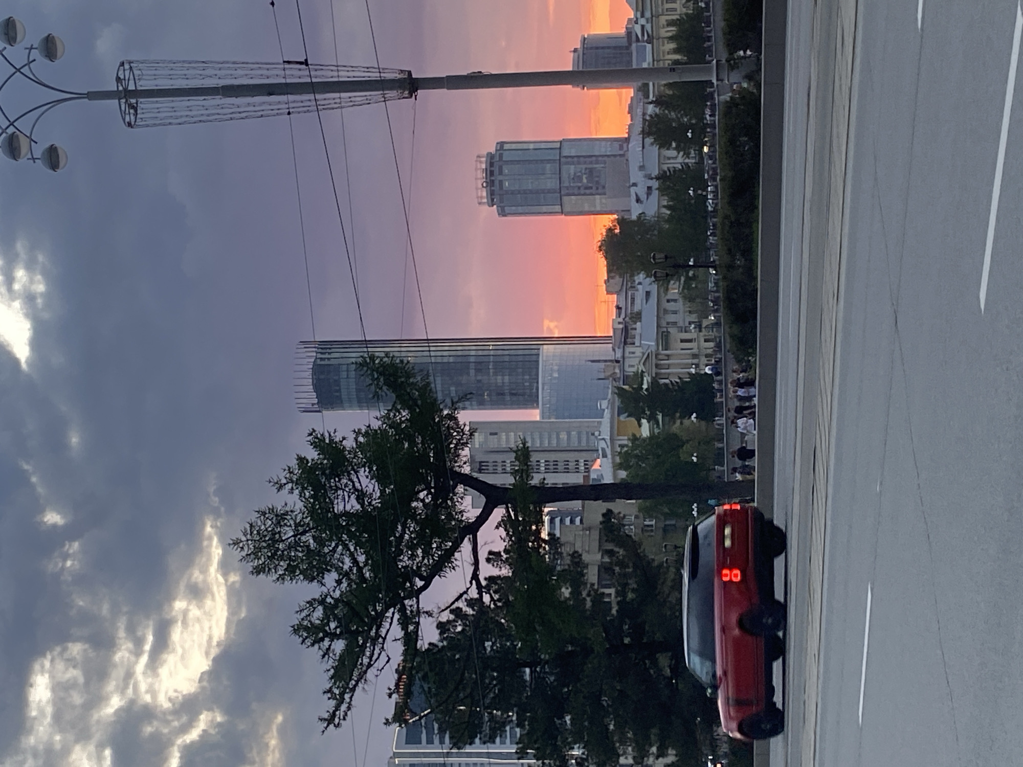 The image captures a street scene in Yekaterinburg, Russia, at sunset. The sky is a blend of purples, grays, and oranges, reflecting the setting sun. Several modern high-rise buildings are visible in the background, silhouetted against the sky. A tree with sparse foliage is positioned on the left side of the frame. A multi-lamp street light is present on the left, with a wire structure. A road is prominent on the right side of the image, with a red car parked on the side. People are gathered along the sidewalk.