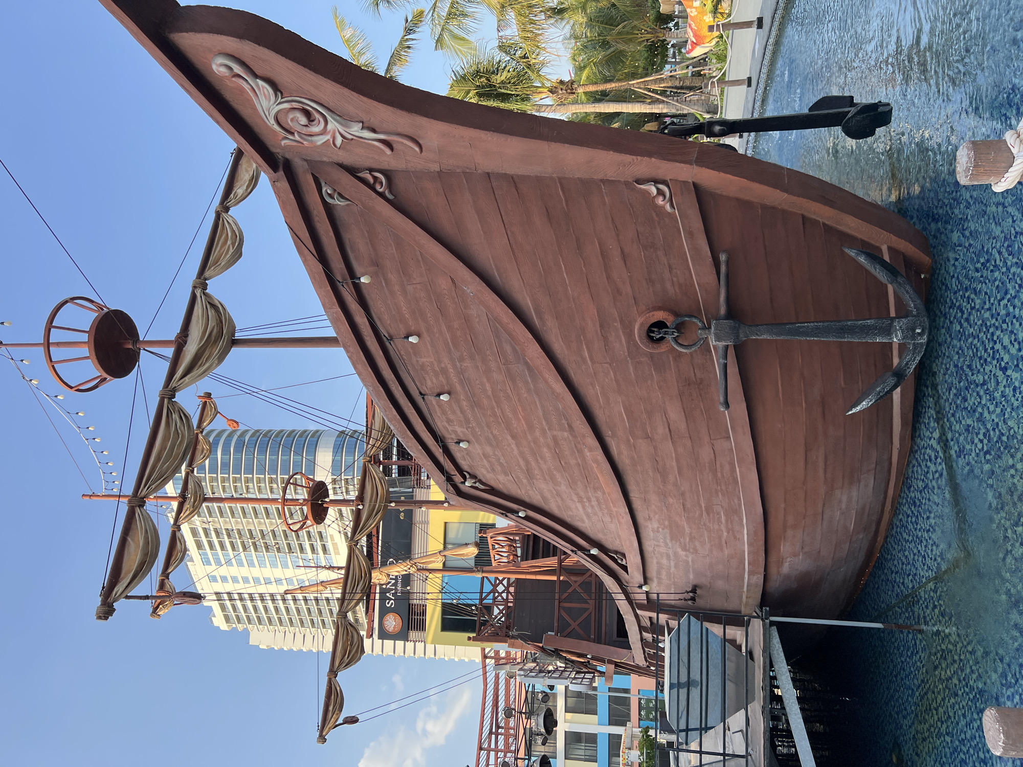 This media file captures a close-up view of a wooden pirate ship, likely a decorative or amusement feature, situated in a pool with blue, patterned tiles. The ship's bow is prominent, showcasing detailed carvings and a large anchor hanging over the side, partially submerged in the water. Multiple furled sails, made of a tan, fabric-like material, are attached to masts. Decorative lights are strung along the ship's structure.
In the background, a modern, multi-story building with a curved, glass facade dominates the skyline, suggesting an urban or resort setting. Palm trees are visible, indicating a tropical climate. The sky is clear and bright blue, suggesting it is daytime, likely with sunny weather.
While no people are directly visible, the presence of the ship and pool implies a recreational area. The scene has a vibrant, possibly tourist-oriented atmosphere, given the juxtaposition of the historical ship design with contemporary architecture. The lighting suggests it is a bright, clear day.
There is visible text on a sign attached to the ship's structure, partially obscured, which reads "SAINT DINING". The overall impression is one of a fun, themed environment in a sunny, tropical location.