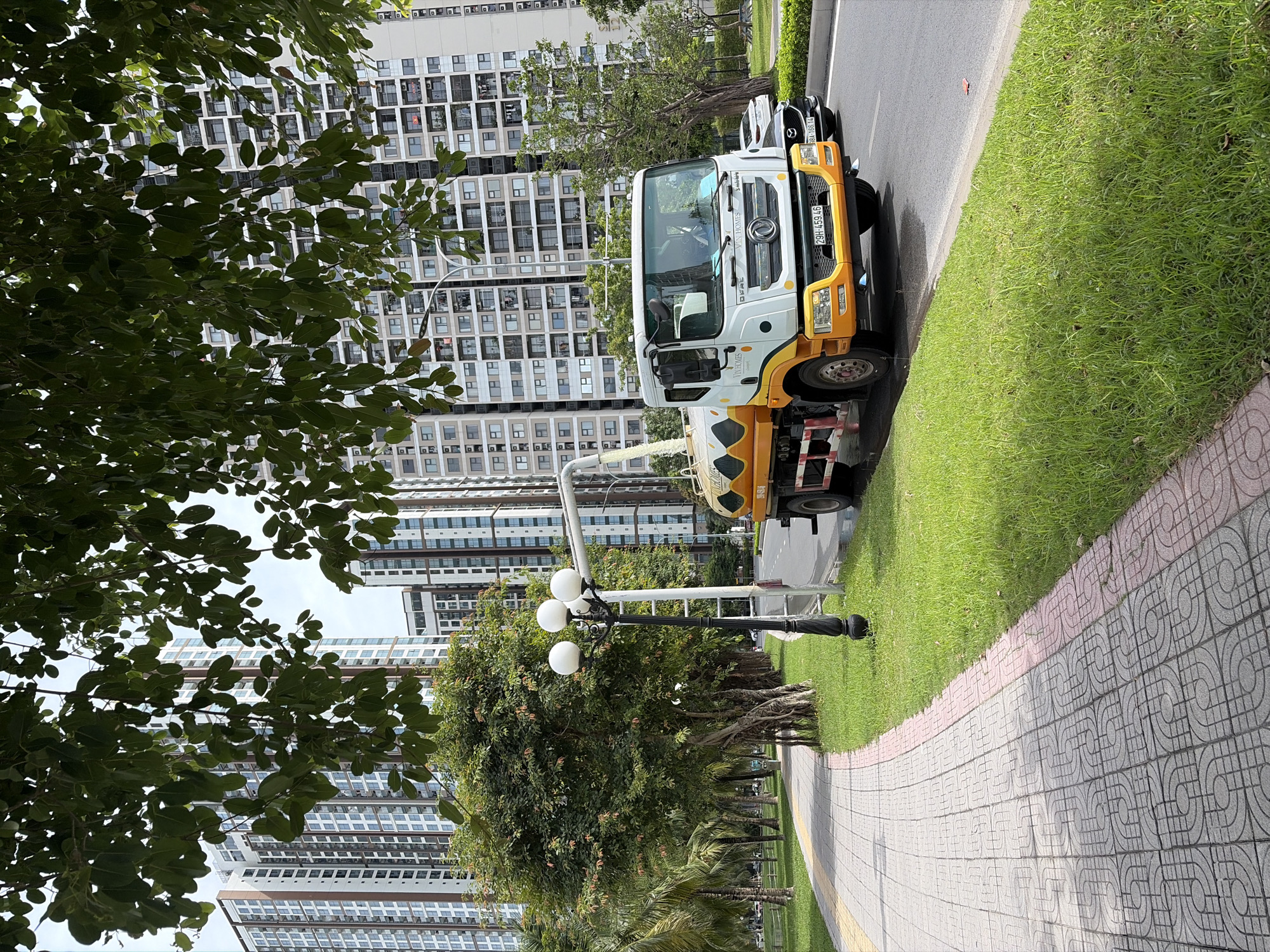In a city environment, likely Hanoi, Vietnam, a white and yellow water truck with the text "VIN HOMES" on its side is parked on the right side of a paved road. The truck has a tank with a wavy orange and white pattern. A white hose extends from the top of the tank and appears to be dispensing liquid. In front of the truck, a black car with a license plate ending in "166.14" is visible.
To the left of the road is a sidewalk paved with grey tiles featuring a circular pattern, bordered by a pink-tiled edge. Adjacent to the sidewalk is a lush green lawn. Further to the left, a row of trees, some with exposed roots, lines the sidewalk. Beyond the trees, a path or park area can be seen, with more palm trees.
In the background, tall, modern apartment buildings with many windows dominate the skyline. These buildings are a mix of light colors, with vertical brown accents. The sky is overcast. A black street lamp with three white globes stands near the sidewalk and the edge of the road. The scene appears to be a well-maintained urban residential area.