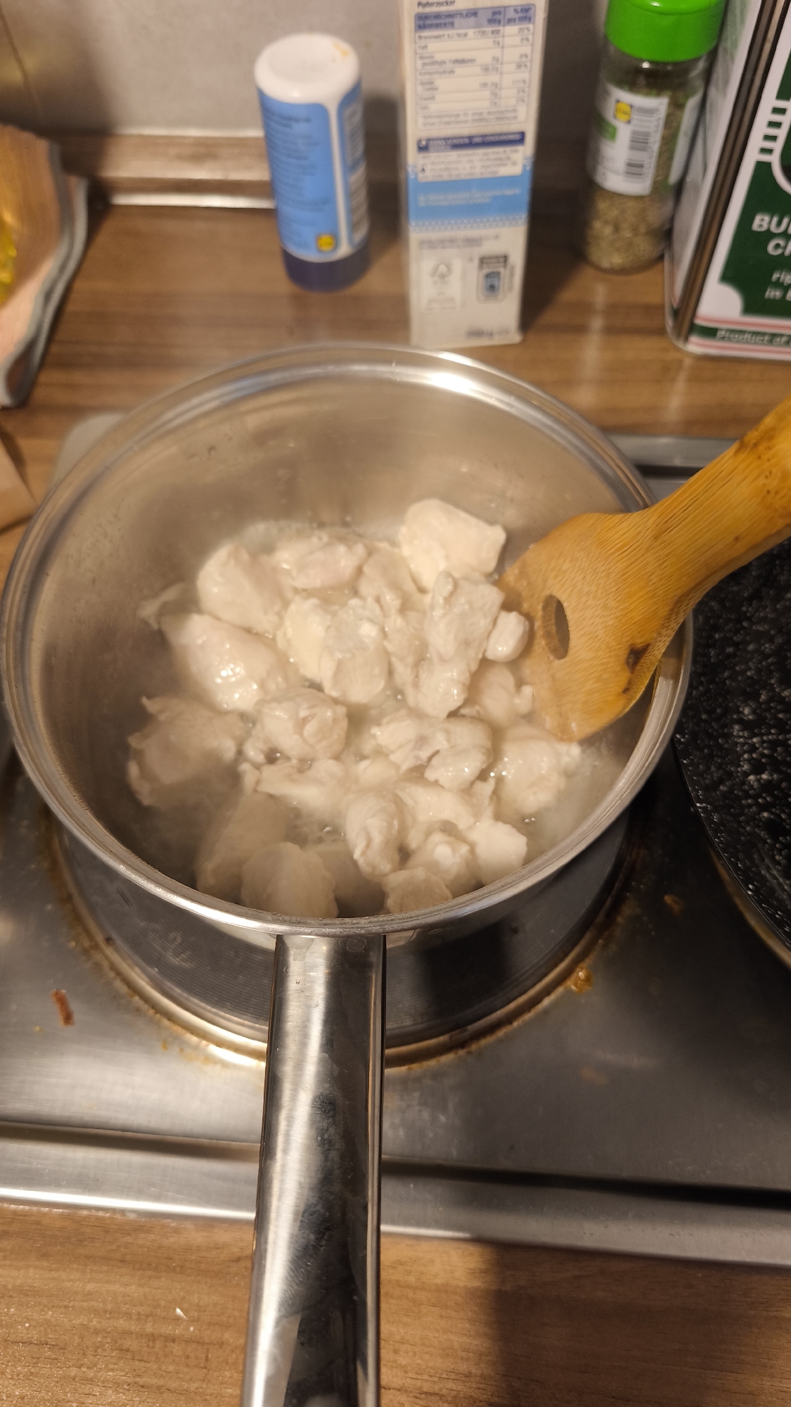 An overhead view captures a cooking scene in a home kitchen. The central focus is a silver pot resting on an electric stovetop, containing numerous diced pieces of white meat, likely chicken, simmering in a light liquid. Steam rises from the pot, indicating heat. A light brown wooden spatula, with a hole in its paddle, is partially submerged in the liquid, stirring or resting among the chicken pieces.
In the background, behind the pot, a wooden countertop is visible. Various kitchen items are arranged on it. To the left, a blue and white shaker-style bottle stands upright. Next to it, a white rectangular carton with nutritional information is visible; parts of the text "Nährwerte" (nutrition facts), "Brennwert" (calorific value), and percentages like "25%" are legible, along with "Butterzucker" (butter sugar). Further to the right, a clear glass spice jar with a green cap, filled with dried green herbs, is present. A larger green-labeled box is partially seen, showing "BUCH CH..." and "Product of" with the country name cut off. A dark, possibly black, pan or cooking surface is partially visible to the right of the main pot. The stovetop surface itself appears metallic with some minor splatters or stains.