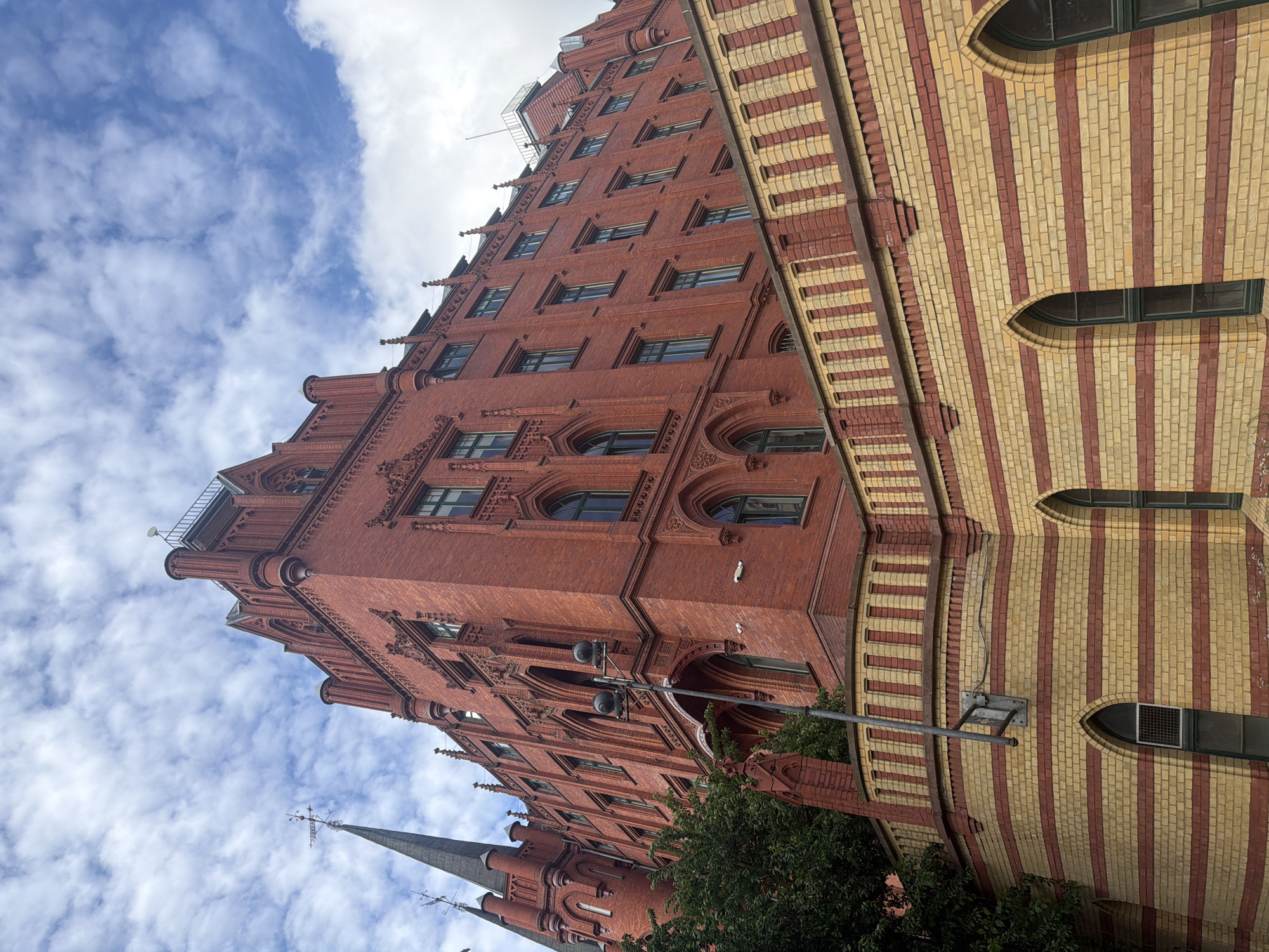 The image shows the historic Red Town Hall building in Szczecin, Poland. The facade is constructed of red brick with gothic architectural elements. There are arched windows with dark frames and decorative brickwork above them. A striped wall made of yellow and red brick is in the foreground, with arched windows at its base. A street lamp is visible attached to a pole extending from this wall. The sky is cloudy with patches of blue.