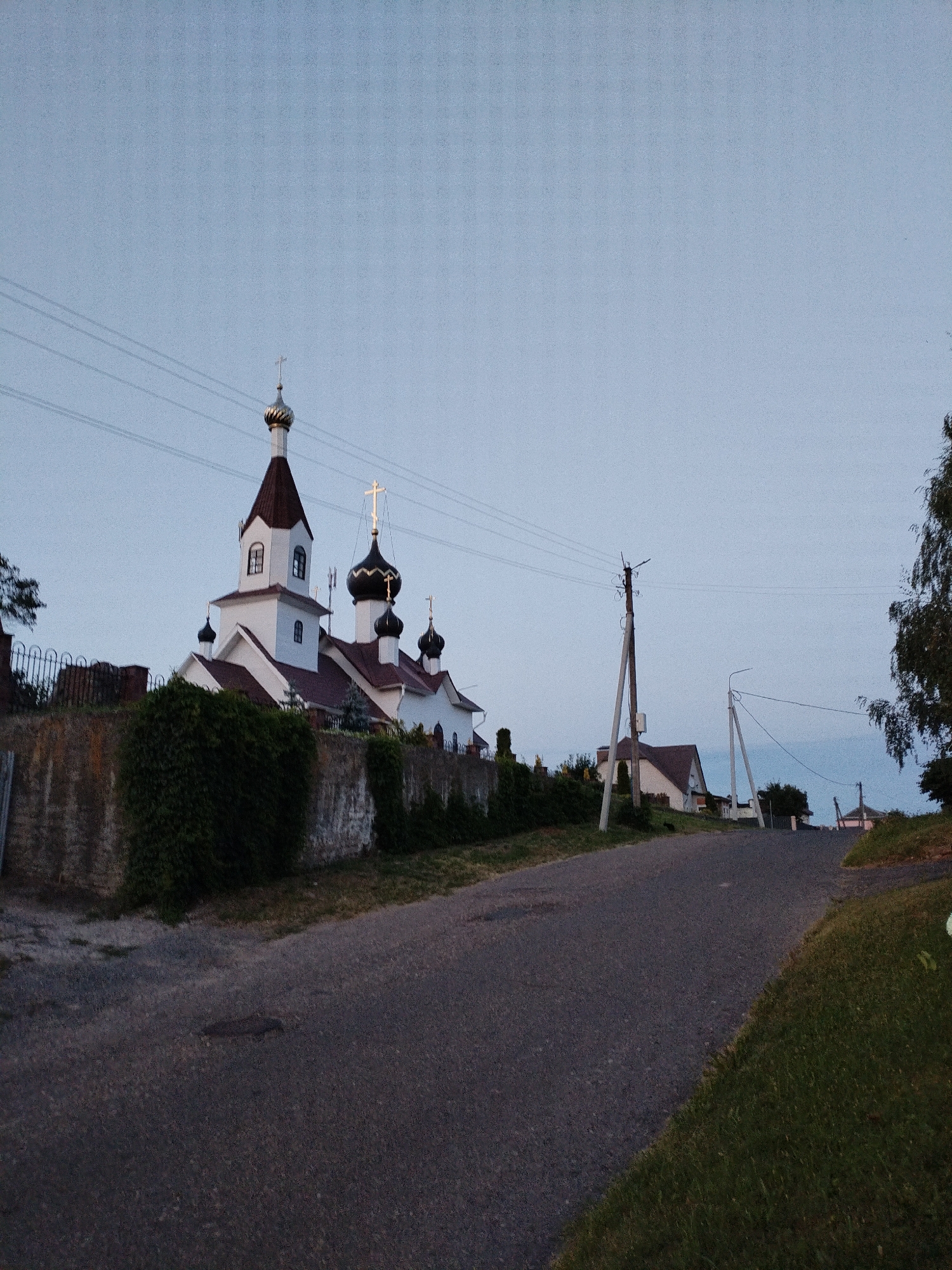 The image depicts a view of an Orthodox church in Bialynichy, Belarus, on a clear day with a pale blue sky. The church, with white walls and reddish-brown roofs, features multiple domes, one of which is topped with a cross. A stone wall, partially covered in ivy, runs along the foreground, separating a paved road from the church property. Utility poles with electrical wires are visible along the road, which slopes upwards towards houses in the distance. Green grass and trees frame the scene. No people are present.