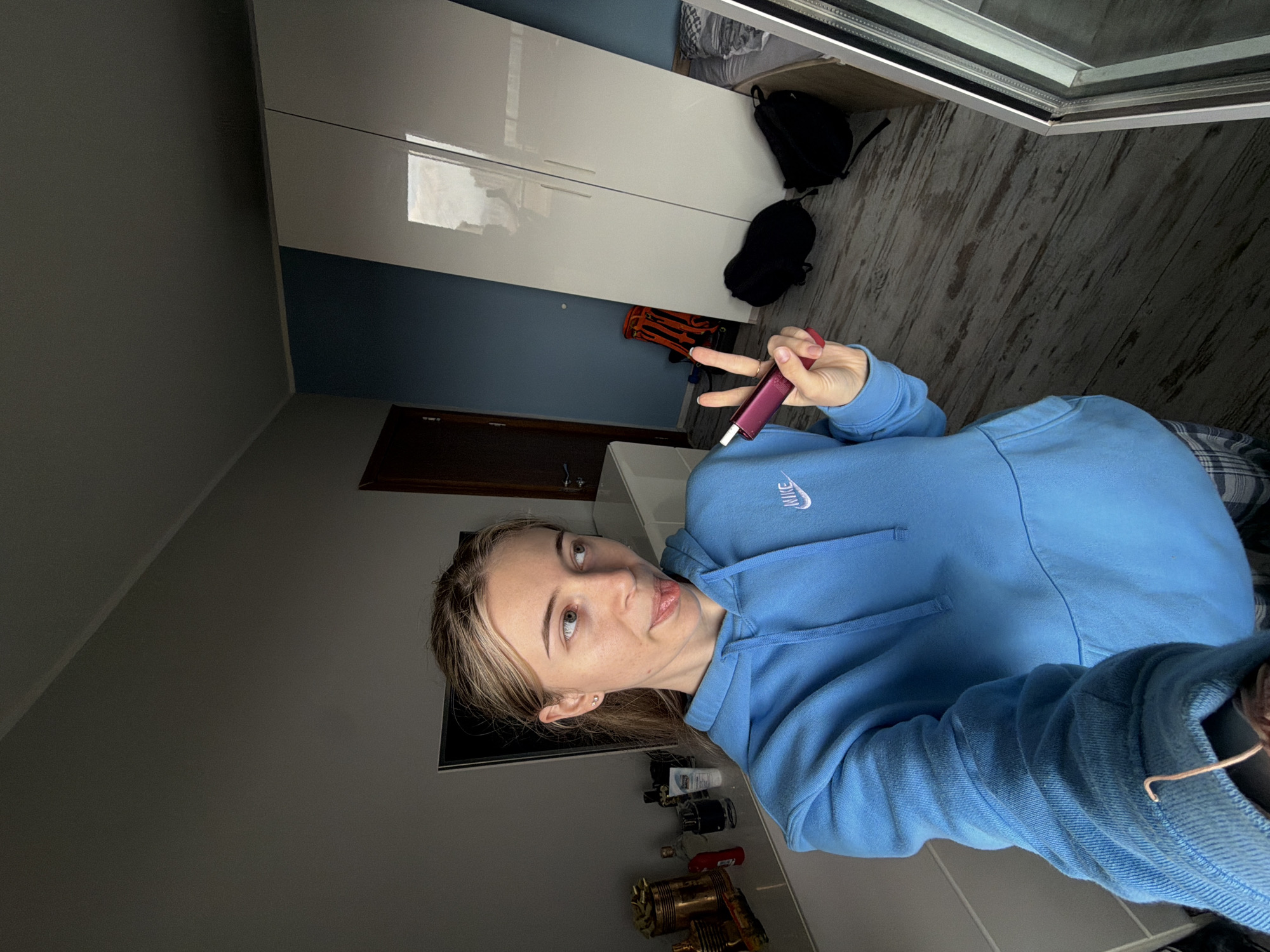 A female individual is captured in a medium shot within a domestic interior in Plovdiv, Bulgaria. She has light brown hair, pulled back, and wears a light blue hooded sweatshirt with a white Nike logo. Her left hand, visible in the lower right quadrant, holds a pink and purple cylindrical device (consistent with a heated tobacco or vaping product) while her index and middle fingers form a "peace" sign. She looks towards the viewer's left with her tongue slightly extended. Plaid pajama bottoms are visible at the bottom of the frame.
The room features a light grey wall to the left and a dark blue wall to the right, meeting a white ceiling. On the left, a white dresser or cabinet is topped with several bottles, metallic containers, and a white tube. A dark, rectangular screen is mounted on the wall above this dresser. To the right, a tall, white, glossy two-door wardrobe stands against the dark blue wall, reflecting the ceiling and possibly a window. On the wooden floorboards, two black backpacks are positioned near the wardrobe. A portion of an orange backpack is visible between the wardrobe and a dark brown wooden door on the far right. A bed with patterned bedding is partially visible in the bottom right corner, adjacent to the wardrobe. The image appears to be a mirror selfie.