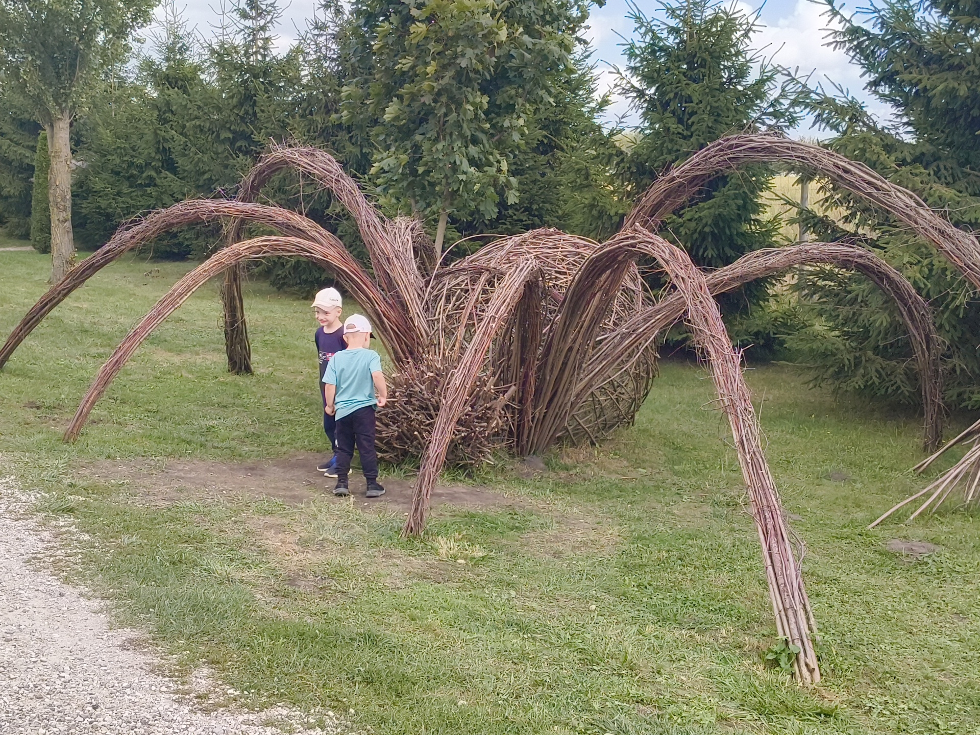 The image depicts an outdoor scene featuring a large, spider-like sculpture made of woven branches, situated on a grassy area. Two young boys, wearing hats, are standing near the sculpture. One boy, in a blue t-shirt, faces away from the camera, while the other, in a dark shirt, is partially visible. The background consists of a dense forest of evergreen and deciduous trees, with a partially visible dirt path in the foreground. The setting appears to be a park or a natural reserve within the Малахавецкі сельскі Савет, Belarus.