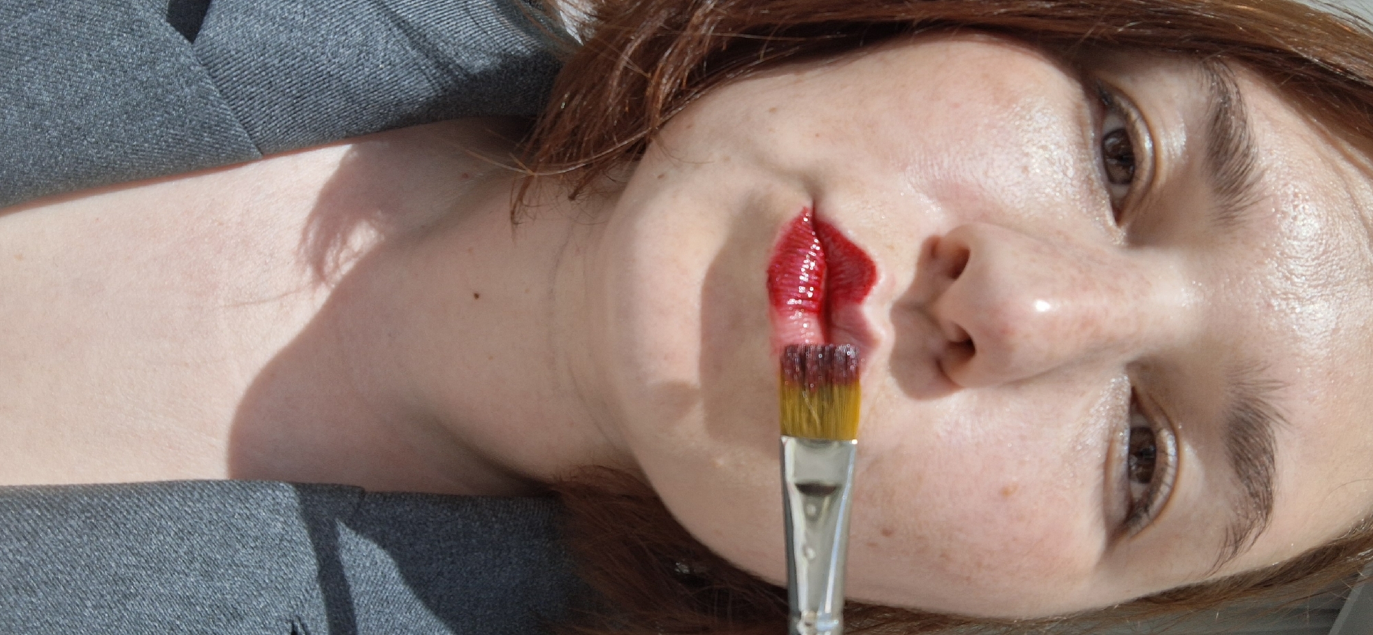 The image shows a woman lying down, angled slightly upwards. She has fair skin with freckles and light brown eyes. Her hair is reddish-brown and appears slightly disheveled. Red paint is applied to her upper lip, smeared upwards towards her nose. A paintbrush with red paint on the bristles is positioned vertically, touching the red paint on her lip. The woman is wearing a grey garment that appears to be a jacket or coat. The background is out of focus, suggesting the photograph was taken with a shallow depth of field. There is no information given in the image to suggest this image was taken in Chaikovsky, Russia.