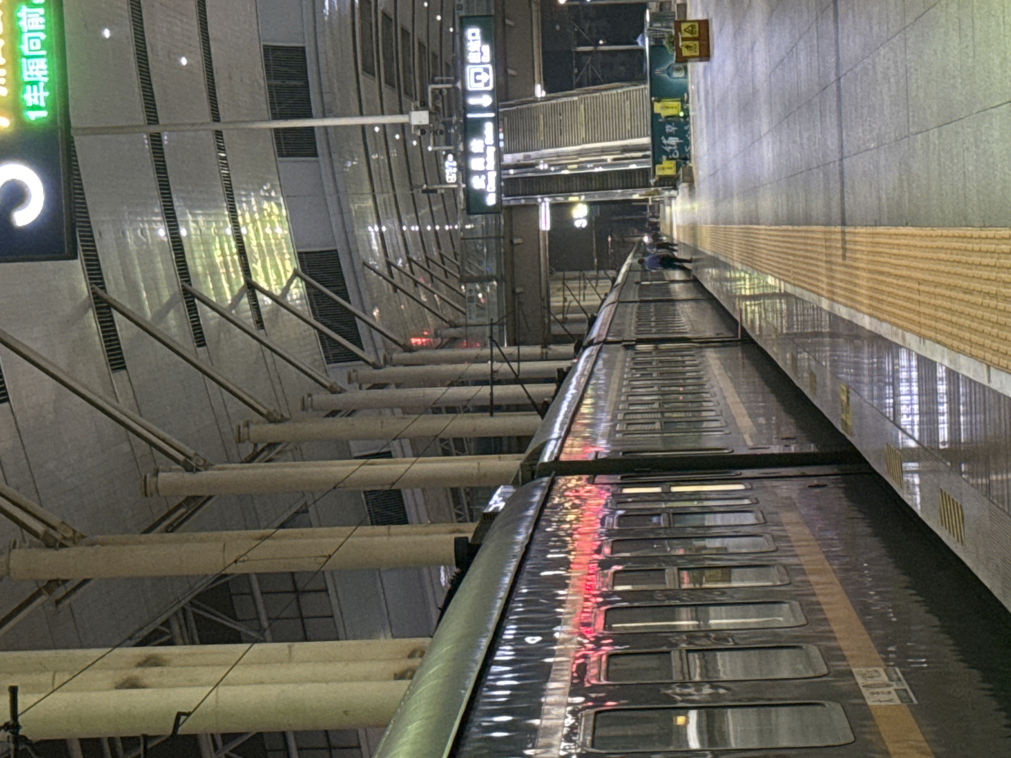 The image shows a train at a station platform. The train is long and dark with reflections on its side. The platform is paved with gray tiles and has a yellow tactile paving strip along the edge. In the background, there are large columns supporting a roof structure. Signs are visible, including one with Chinese characters and a number "3". An escalator is also visible in the background. A few people are walking on the platform. The scene appears to be indoors at night or in low light conditions. The location is Wuchang Railway Station in Wuchang District, China.