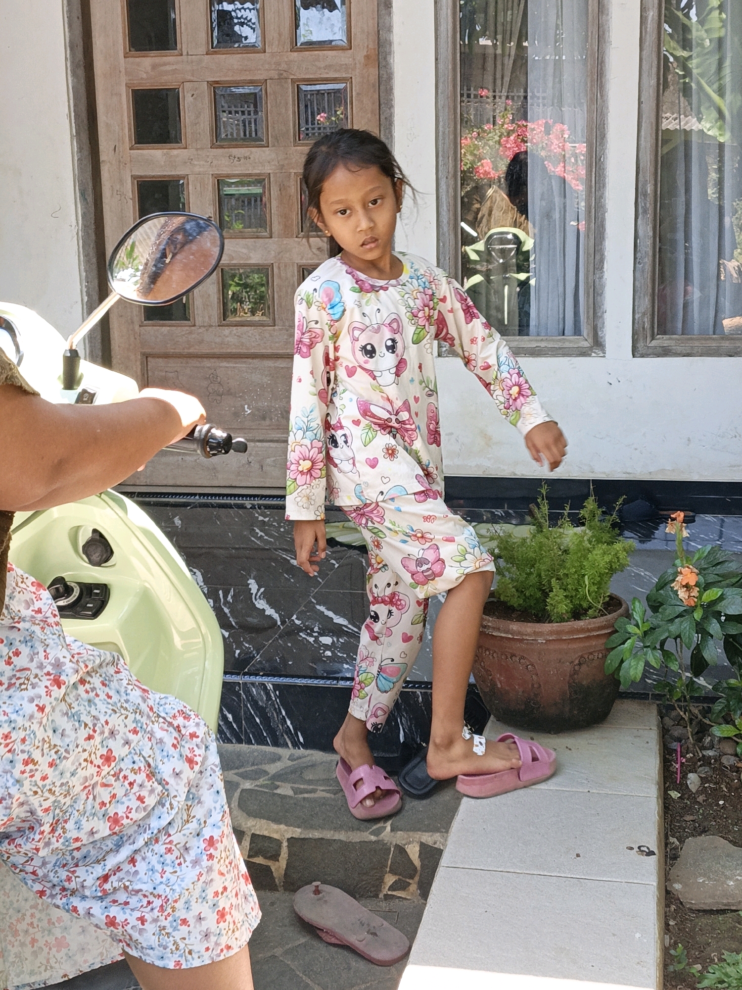 A young girl in pajamas with a cartoon character print stands on a tiled porch in front of a wooden door. She is wearing pink slides on her feet. To her left, a portion of a light green scooter is visible, with a person's arm resting on the handlebars. Next to the scooter, another person is seated, wearing a white dress with a floral pattern. A potted plant sits to the right of the girl. The scene appears to be outdoors, in front of a residential building. The location is identified as Cikal, Indonesia.