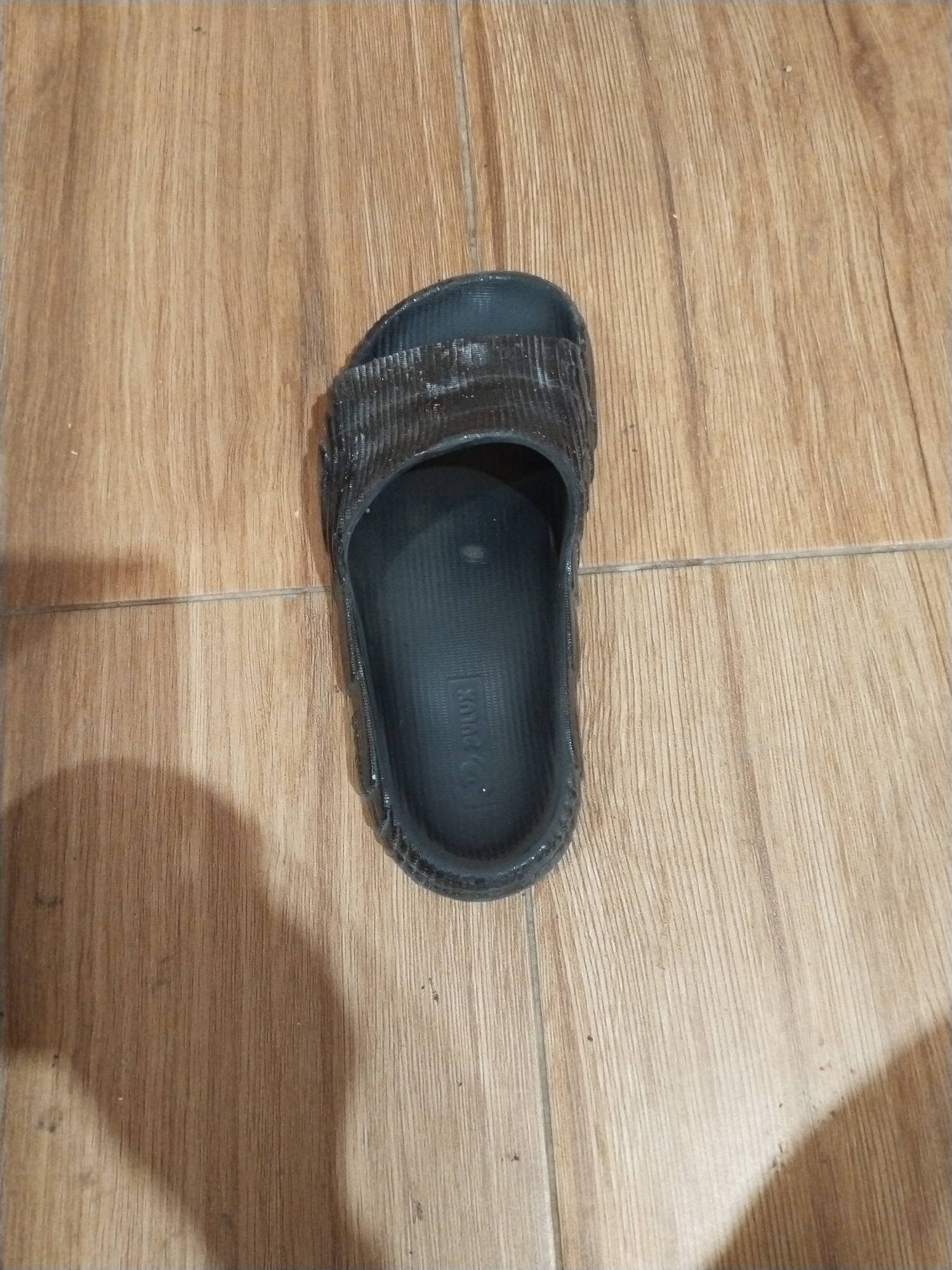 A single, black, rubber flip-flop lies on a light brown tiled floor. The flip-flop is positioned in the center of the frame, with its open toe facing upwards and its heel towards the bottom. The surface of the flip-flop is textured with horizontal ridges on the strap and a smooth, molded footbed. A faint shadow of the flip-flop is visible to its left, suggesting an overhead light source. The floor tiles are arranged in a grid pattern and show wood-grain detailing. The scene appears to be indoors, likely in a residential setting within the city of Prigi, Indonesia. There are no people or other objects visible in the image.