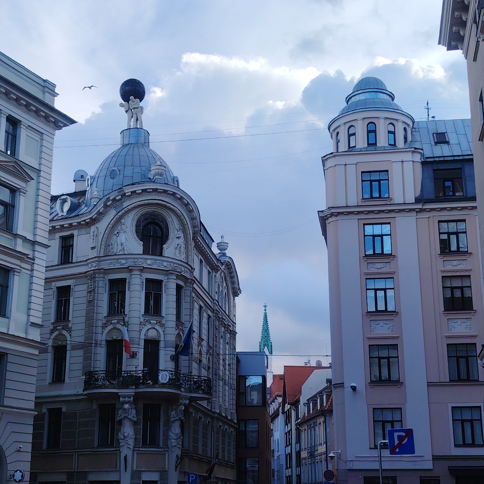 The image shows a street scene in Riga, Latvia, with several historic buildings. On the left, a light-colored building with ornate architectural details features a dome topped with a sculpture of three figures holding up a globe. A bird is seen in flight in the sky above this building. To the right, another building has a rounded turret with arched windows and a domed roof. The sky is partly cloudy with a pale blue hue. A green spire of a distant church or cathedral pierces the skyline between the buildings. The buildings have multiple windows, some with reflections of the sky. The overall impression is of an urban environment with well-preserved historical architecture.