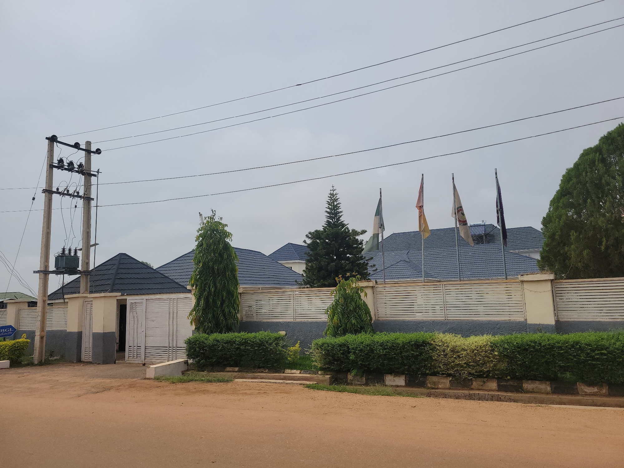 The image depicts the exterior of a building in the city of Barakin Akawo, Nigeria. A prominent feature is a utility pole with electrical equipment, including a transformer, situated on the left side of the frame. The building itself has a modern design with dark grey, tiled roofs and white horizontal slatted fencing. Several flags are visible flying on poles in front of the building, adding a touch of formality. The grounds are landscaped with manicured green bushes and trees, suggesting a well-maintained property. The road in the foreground is unpaved and appears to be made of red earth. The sky is overcast, indicating a cloudy day. There are no people visible in the image.