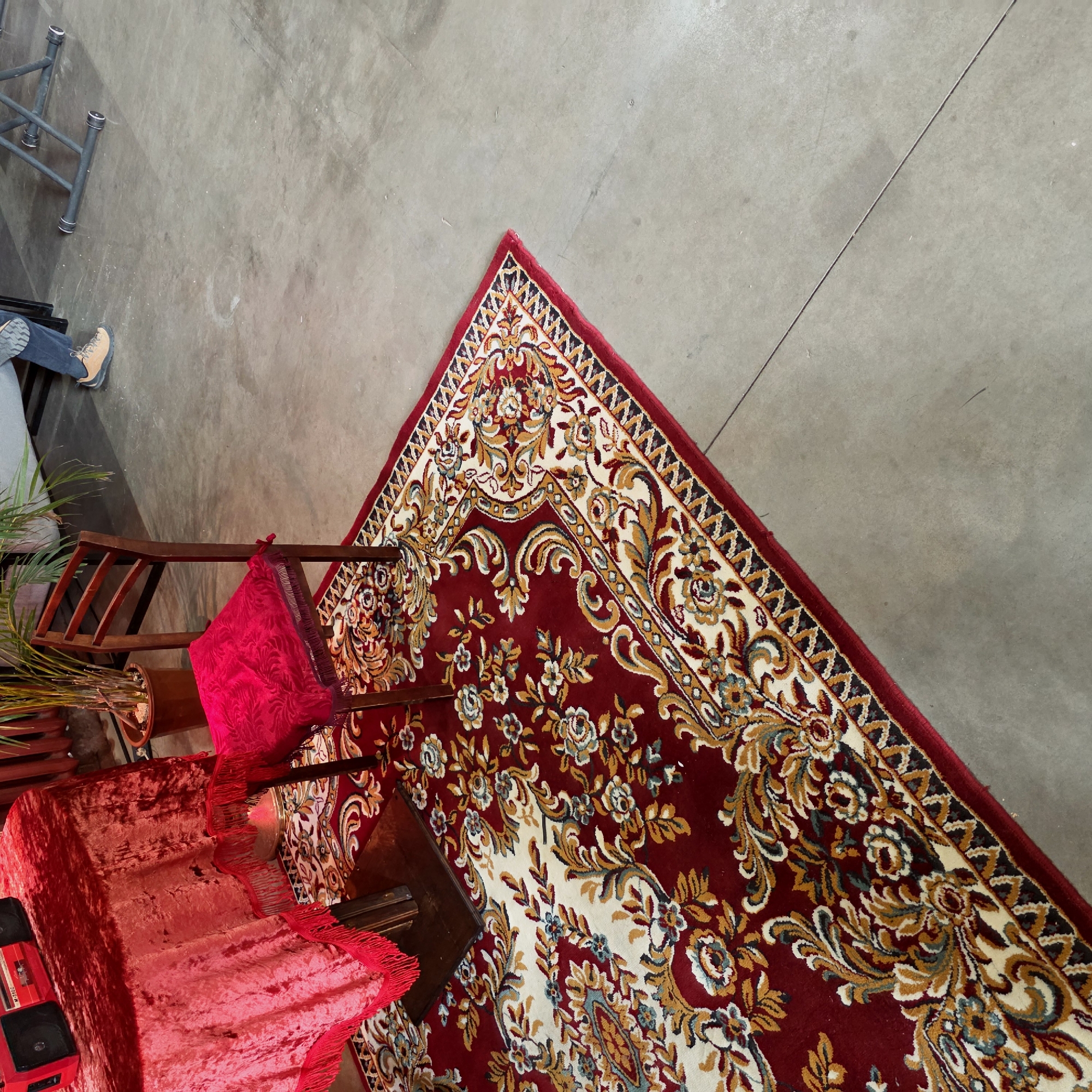The scene presents an overhead view of a section within what appears to be a commercial or residential building, potentially located in Saint Petersburg, Russia, given the typical architectural styles and interior design choices found in the region. A portion of a patterned rug in predominantly red, gold, and ivory tones sits on a gray concrete floor. The rug features ornate floral designs and is bordered by a thick red trim with geometric patterns. Adjacent to the rug is a set of dark wood chairs, one of which is draped with a bright red fabric adorned with fringed edges. A staircase-like structure is covered with the same red fabric, also fringed. In the upper-left corner, there's a glimpse of a metal scaffolding structure, and part of a person's lower body, including a shoe and blue jeans, is visible. Additionally, there's a potted plant with slender green leaves, partially obscured by the chairs. The overall lighting in the scene is somewhat diffused, creating soft shadows and muted colors.