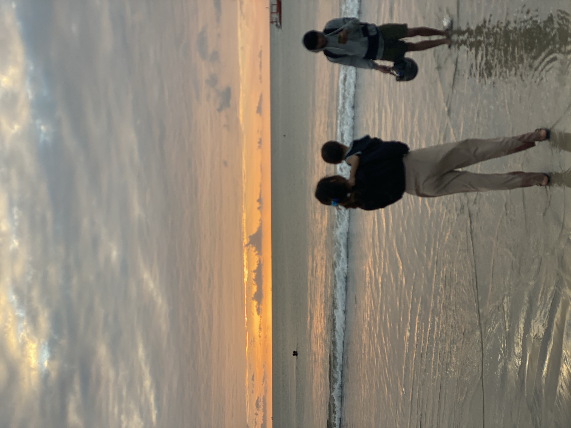 A wide shot of a beach at sunset in Kuta Selatan, Indonesia, shows three figures silhouetted against the orange glow of the horizon. The sky is filled with soft, textured clouds in shades of gray and pink, with the sun's light creating a warm gradient. The ocean is calm, with gentle waves lapping at the shore, reflecting the colors of the sky.
In the foreground, a woman stands facing away from the camera, holding a small child. The woman is wearing a dark top and light-colored pants. To the right, a man stands in the shallow water, holding a helmet and looking at his phone. He is wearing a light gray hooded jacket and dark shorts. In the distance, a small red boat is visible on the water. Two figures are also in the water further out, appearing to be swimming or wading. The sand is wet and reflects the light, creating a shimmering effect. The overall scene depicts a serene evening at the beach.