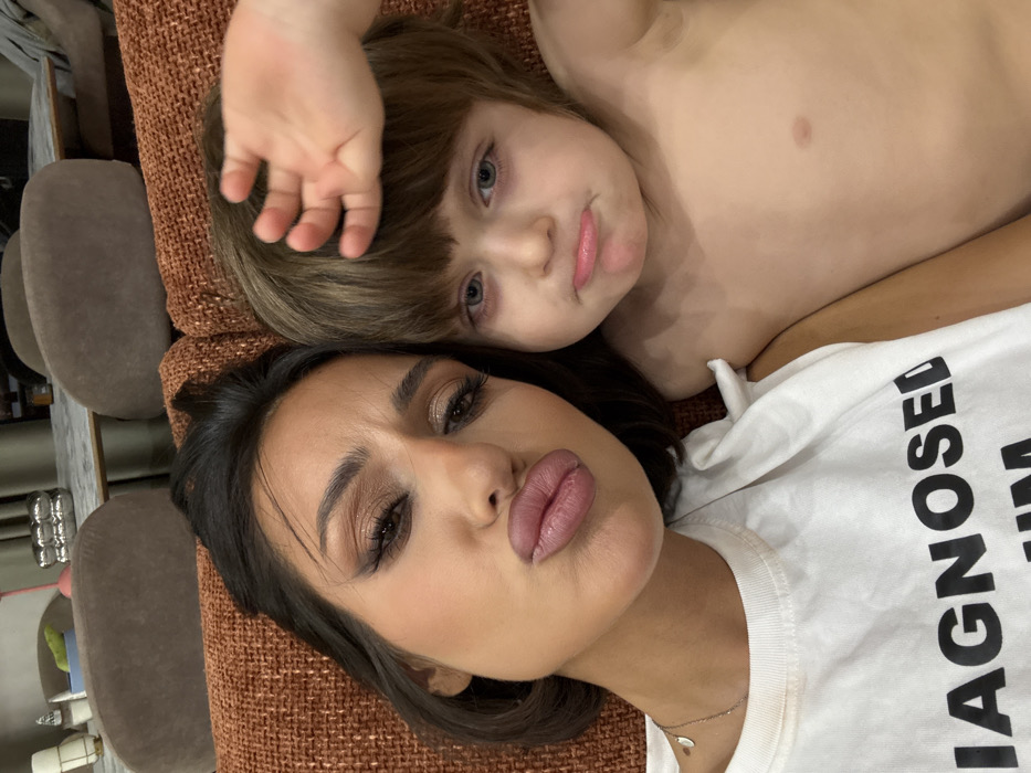 A selfie-style image features a woman and a child, likely a mother and son, lying on a brown textured surface, possibly a couch or bed. The woman, in a white t-shirt with bold text, faces the camera with pursed lips. The child, with brown hair and light eyes, is above her. Both are positioned close together, creating a cozy, intimate atmosphere.