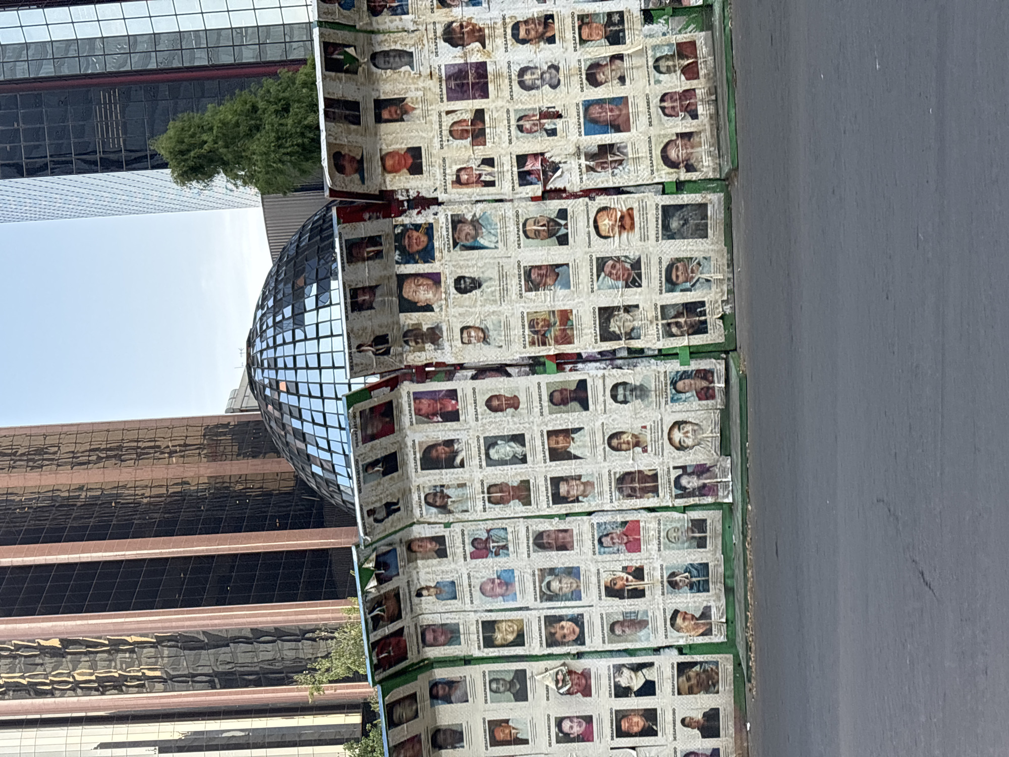 This media file captures a powerful street art installation or memorial in an urban setting during bright daylight. The main subject is a large, multi-panel display, oriented vertically, covered with numerous individual photographs of people. Many of these photos are accompanied by the Spanish word "DESAPARECIDO" (disappeared, male) or "DESAPARECIDA" (disappeared, female), indicating that the display is dedicated to missing persons. The photos are arranged in a grid pattern, creating a dense mosaic of faces, and some show signs of wear.
In the background, to the left, a large, spherical structure covered in mirrored tiles is visible, reflecting the surrounding environment. Beyond this sphere and the display, modern, tall buildings with glass and dark facades rise against a clear, bright sky, suggesting a bustling city environment. Green foliage from trees is visible between the buildings and the display. The foreground, to the right of the display, shows a plain, grey paved surface, likely a road or a wide sidewalk. The overall impression is one of solemn remembrance and a public plea for awareness regarding the disappeared.