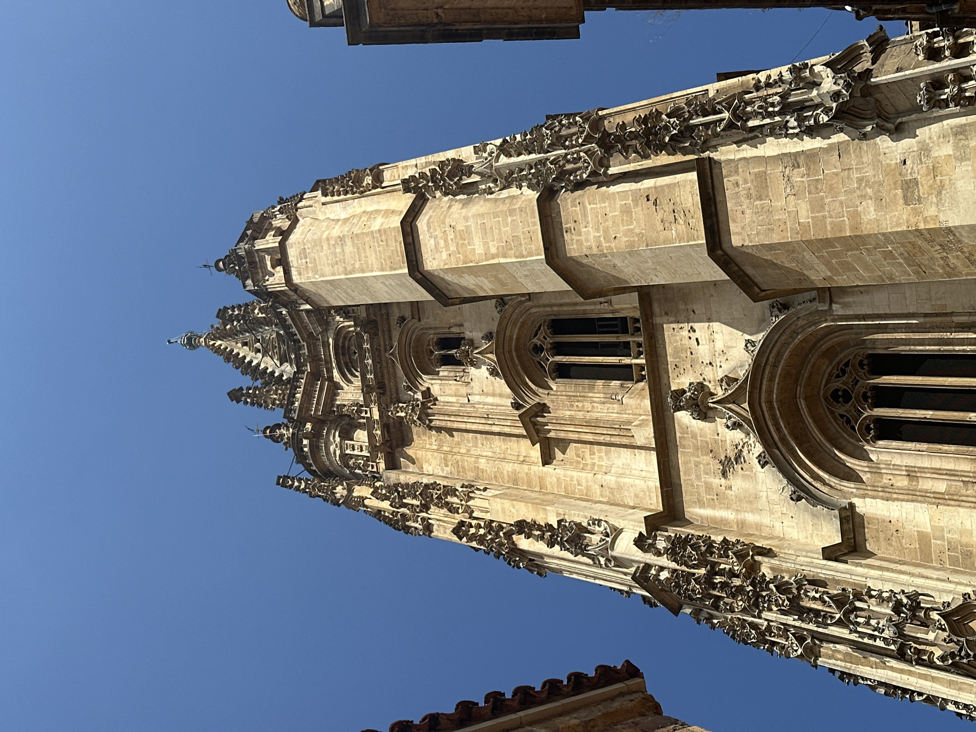 A low-angle, close-up shot captures the intricate stonework of the bell tower of Oviedo Cathedral in Oviedo, Spain. The bell tower is constructed of light-colored stone blocks and features ornate Gothic architecture, including pointed arches, delicate tracery, and numerous small spires and decorative elements. Tall, narrow windows with pointed tops punctuate the tower. The sky above is a clear, deep blue. To the left and right of the tower, partial views of other buildings are visible, one with a reddish-tiled roof and the other appearing to be a darker, more modern structure. The lighting suggests it is daytime with the sun casting shadows on the tower's features.