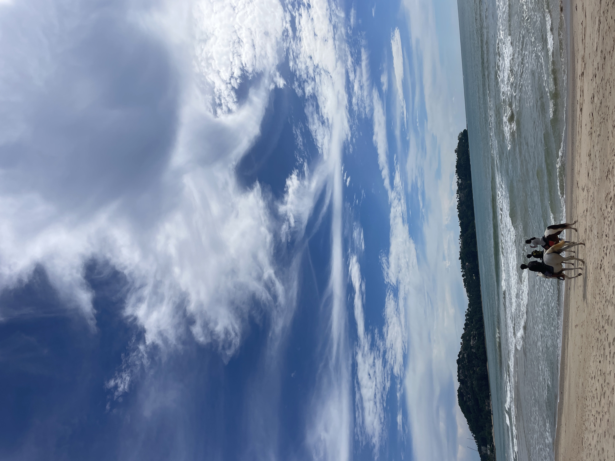 In Choeng Thale, Thailand, on a sandy beach, three individuals are riding horses near the shoreline. The ocean is calm with small waves, and a green, tree-covered island with buildings on it is visible in the distance. The sky is a vibrant blue, filled with scattered white clouds of various sizes and shapes. The overall scene depicts a serene beach activity under a partly cloudy sky.