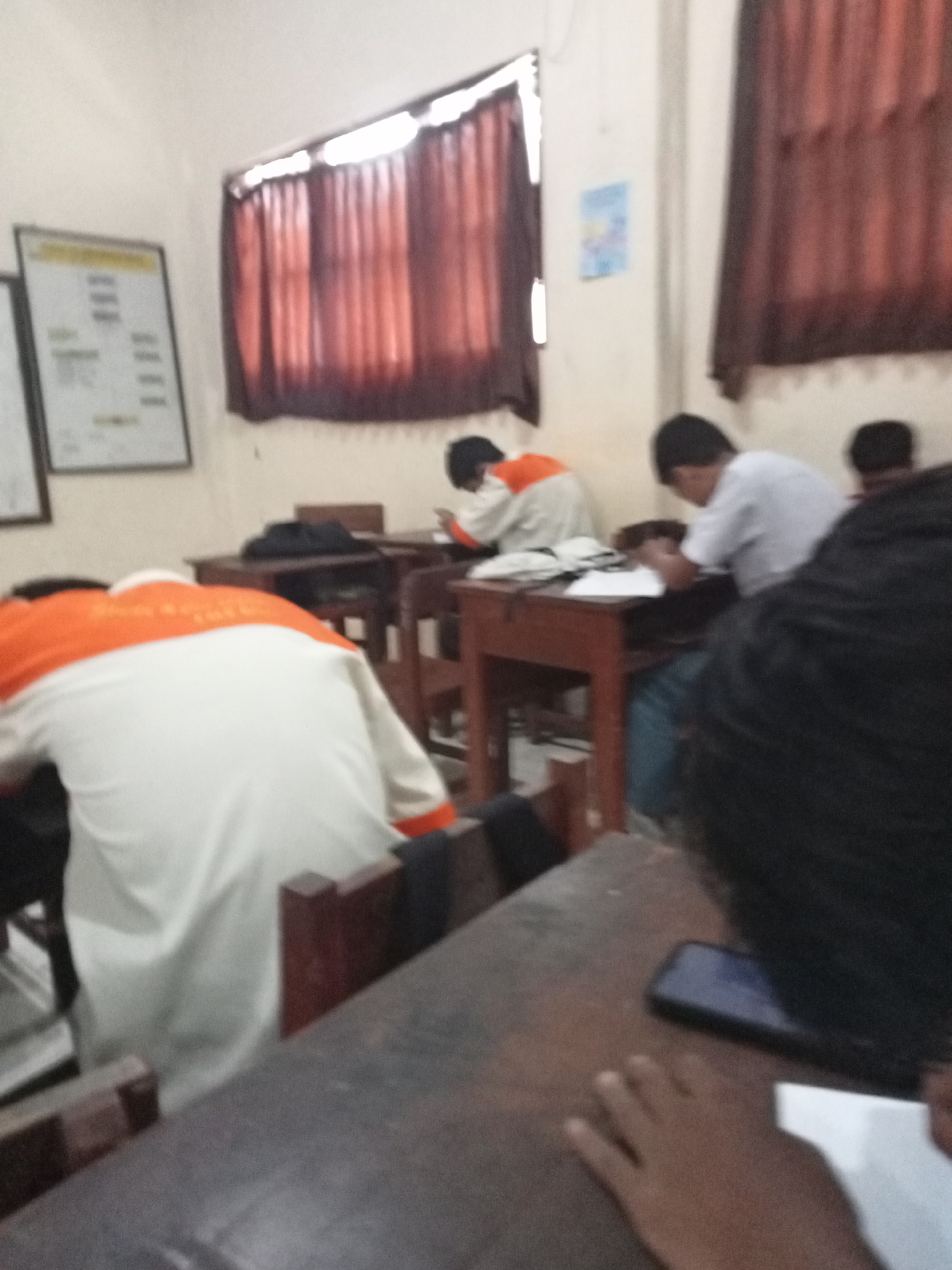 The image depicts an indoor scene, likely a classroom, with several individuals present. The environment appears to be a school in Secang, Indonesia, indicated by the presence of posters on the wall and the overall classroom setup.
In the foreground, a person's back is visible, wearing a white shirt with orange accents. This individual is leaning forward, with their head obscured. To the right, another person in a white shirt is seated at a desk, looking down at their hands, which are resting on a surface likely containing papers or books. Further to the right, a third individual is partially visible, also seated.
In the background, a poster or notice board is mounted on the wall. Red curtains hang over a window, suggesting a typical classroom window treatment. The desks are made of wood, and the lighting appears to be artificial, casting a warm glow on the scene. The overall impression is one of a quiet, possibly study-focused environment.