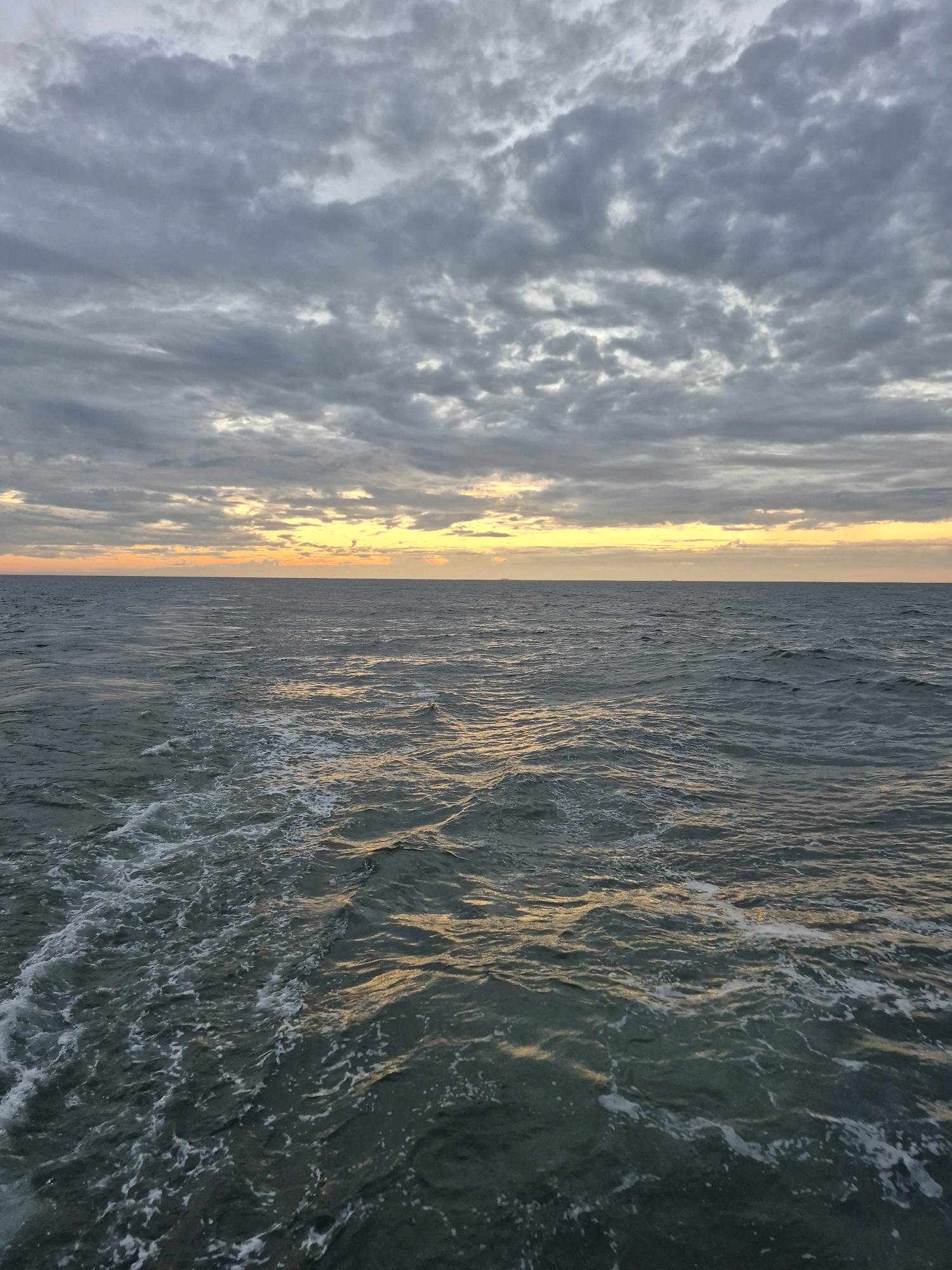 A wide shot captures a cloudy sky over a calm ocean at dawn or dusk. The sky is filled with layers of gray clouds, with a sliver of yellow and orange light breaking through at the horizon. The ocean is dark blue and appears to be moving, with small waves creating white foam on the surface. A wake is visible on the left side of the image, suggesting a boat or ship has recently passed through. The scene is serene and vast, with no visible land or structures in the distance, creating a sense of isolation and natural beauty. The location is Unknown, Germany.