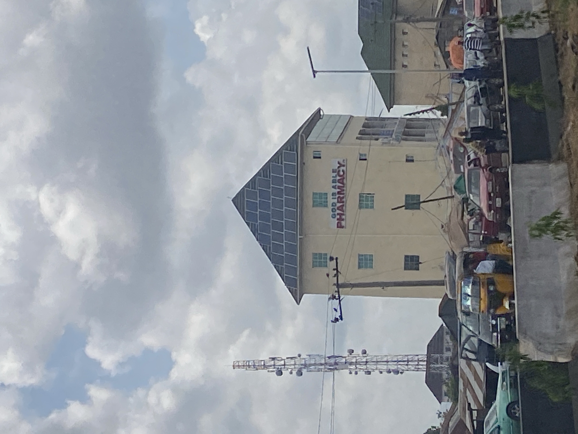 In Ado, Nigeria, a multi-story building with a pointed roof adorned with solar panels stands prominently. A banner on the building reads "GOD IS ABLE PHARMACY." To the left, a tall telecommunications tower rises against a cloudy sky. Below, a street scene unfolds with various vehicles, including cars, a tricycle, and a van. People are visible near the vehicles and walking on the street. The environment appears to be an urban setting with the presence of street vendors and makeshift stalls.