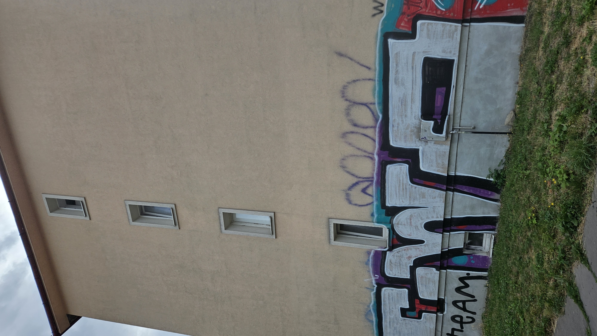 The image shows the side of a pale yellow building with a red-tiled roof. The building has multiple narrow windows arranged vertically. The lower section of the wall is covered in graffiti, with large white, purple, and black lettering dominating the lower half of the frame. The words "ENT" are clearly legible in the graffiti. To the left of this, the word "team" is also visible in black lettering. Above the large graffiti, there are faint purple spray-painted shapes that appear abstract or illegible. The building is situated next to a patch of green grass with some yellow wildflowers and a paved walkway. The sky above is overcast. The scene is consistent with an urban environment in Prešov, Slovakia, likely an apartment building or commercial structure.