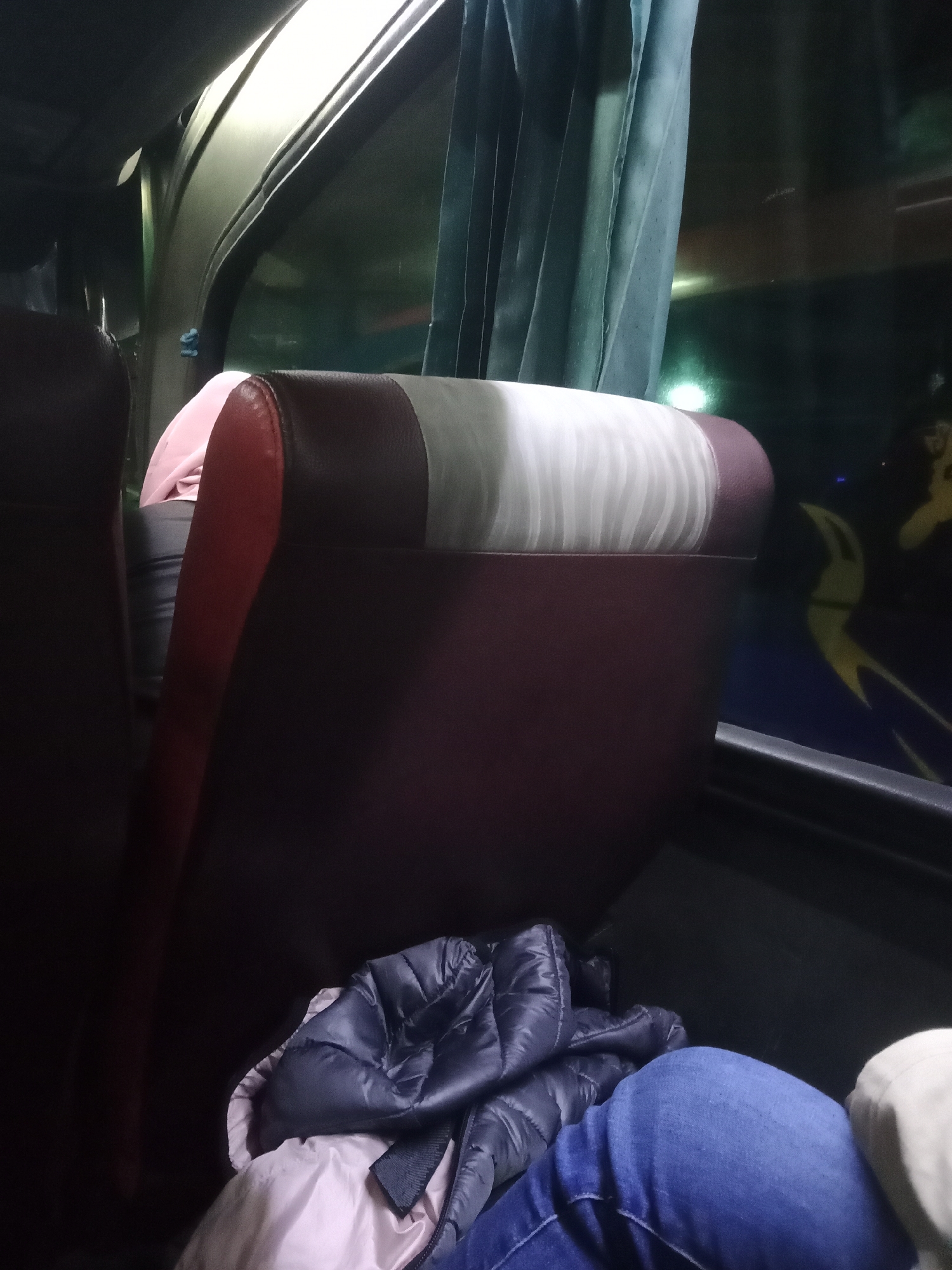 The image depicts the interior of a bus, likely during nighttime travel, as evidenced by the darkness outside the window and the interior lighting. The focus is on a row of seats, with the foreground occupied by a blue padded jacket and the legs of a person wearing blue jeans. The seats are upholstered in a dark red material, with a lighter section at the headrest featuring a striped pattern in white and gray. A pink garment is visible between the seats. Outside the window, a blurred yellow graphic is visible on the exterior of another bus, suggesting the presence of multiple vehicles. The overall scene suggests a journey, possibly a long-distance one, with passengers resting or preparing to rest. The location in Surakarta, Indonesia is not explicitly evident from the visual information in the image.