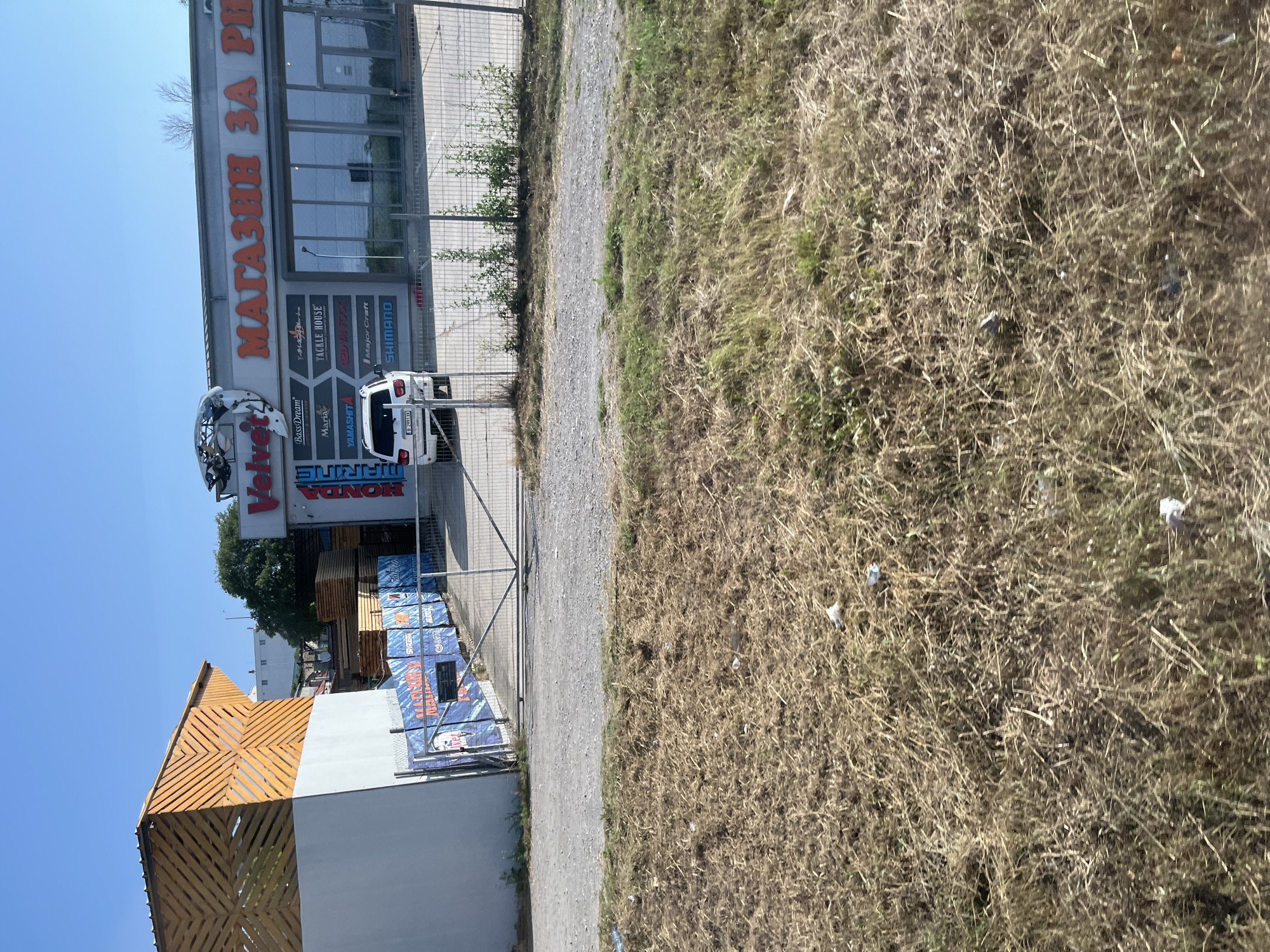 The image shows the exterior of a fishing tackle shop called "Velvet" located in Varna, Bulgaria. The shop's facade features large orange lettering spelling "МАГАЗИН ЗА РИБ" (which translates to "Fishing Shop"). A white SUV is parked in front of the shop. The shop is situated next to a building with a wooden, geometric facade and a fence encloses the area in front of the store. In the foreground, dry, overgrown grass and some scattered litter are visible. The sky is clear and blue, indicating a sunny day.