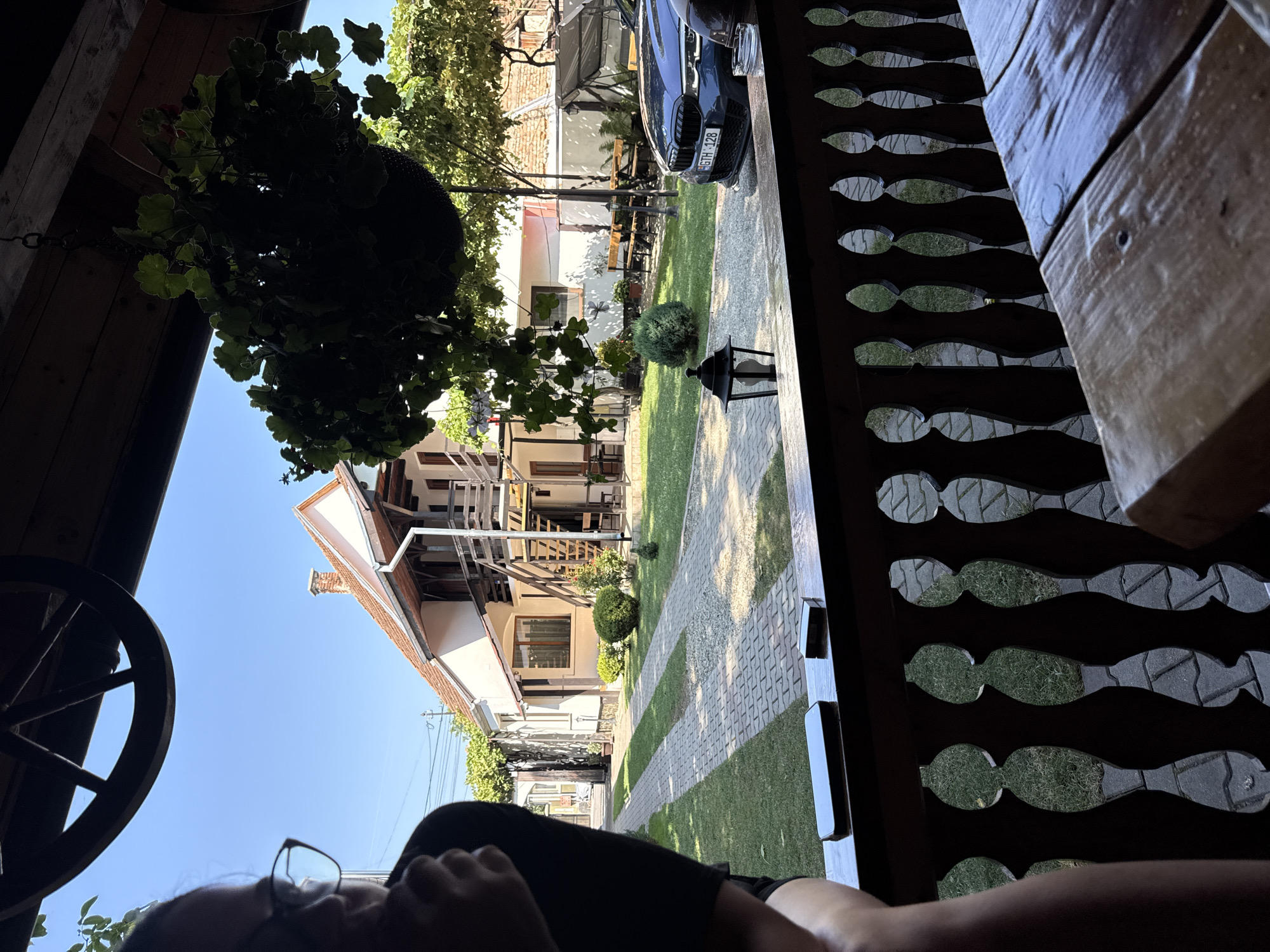 The image shows a view from a wooden porch in Cârțișoara, Romania. In the foreground, a wooden railing and a table are visible. Beyond the railing, a well-maintained yard with green grass and paved pathways leads to a two-story house with white walls and a red tile roof. A dark blue car with a license plate reading "GTH 128" is parked in the yard. A hanging basket of green plants is suspended from the porch roof. The sky is clear and blue. A person wearing glasses is partially visible on the left side of the image.