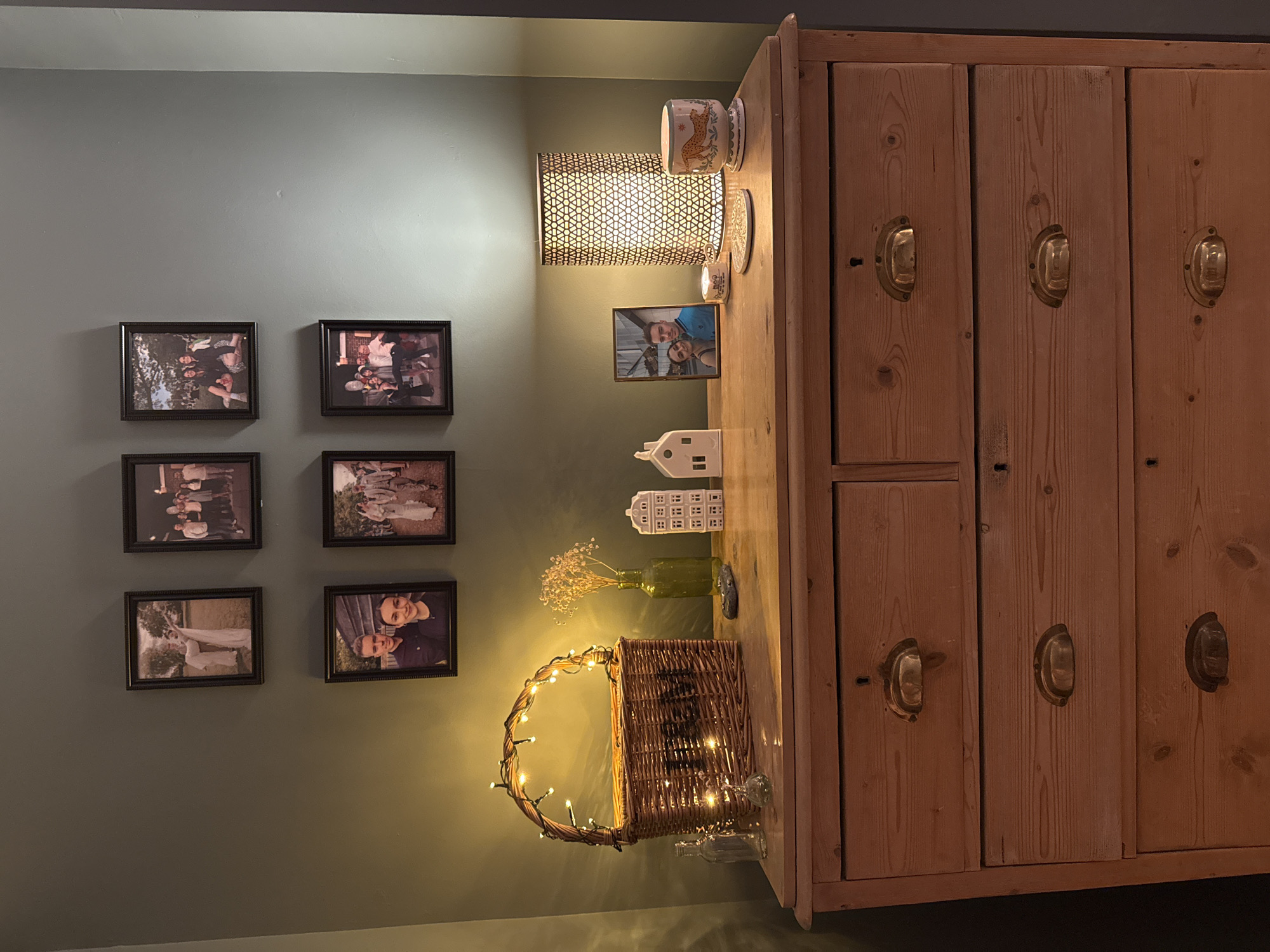A wood dresser is positioned to the right of the frame. On top of the dresser, there is a lamp, picture frames, decorative houses, flowers in a vase, and a basket wrapped in lights. To the left of the dresser, there is a wall with six framed photographs arranged in two rows of three. The walls are painted a dark green color. The scene appears to be indoors. This location is not specified in the image, only the county.