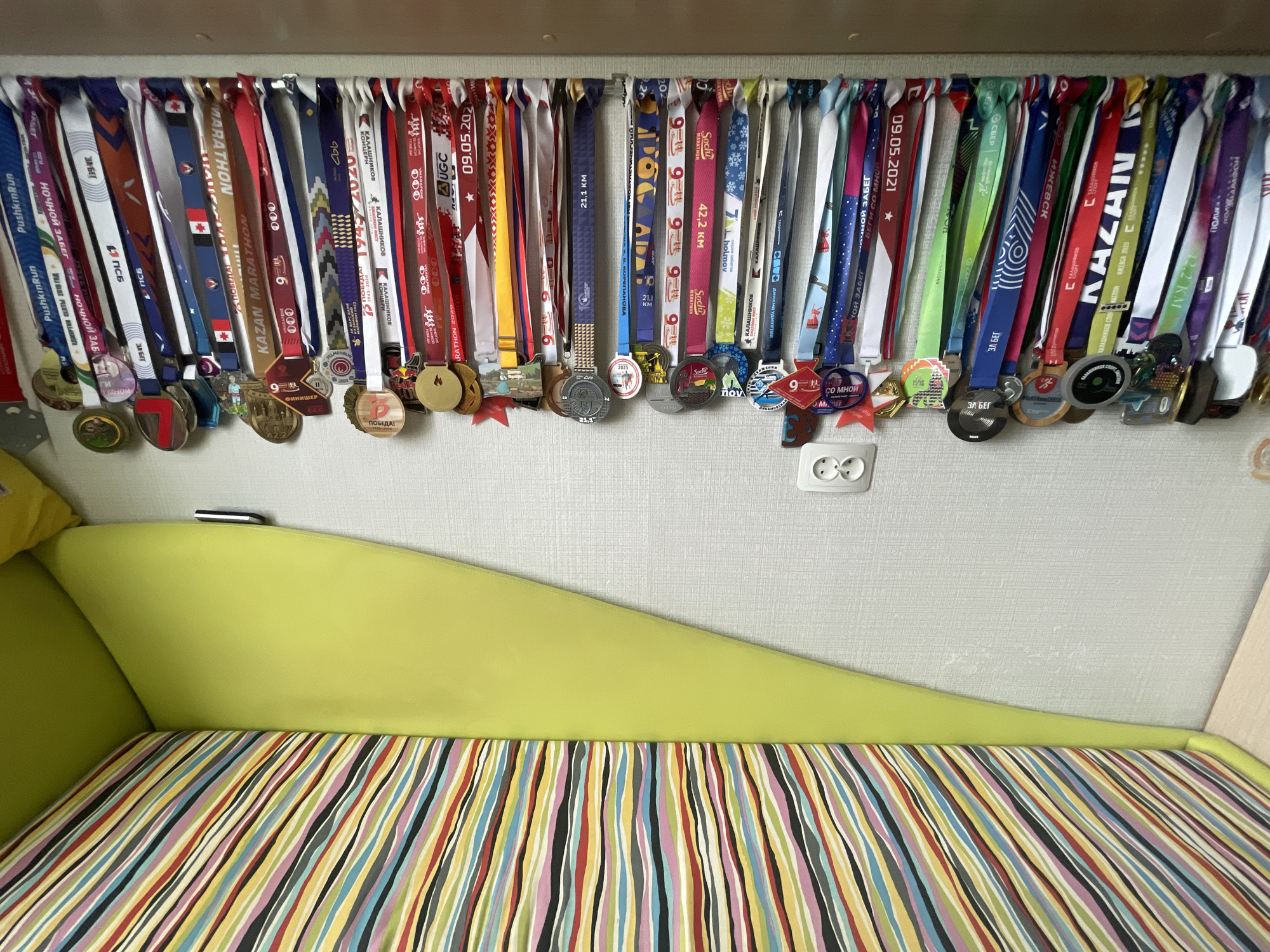 A vibrant collection of approximately 40-50 running medals is displayed, hanging from their colorful ribbons on a white rail against a light-colored, textured wall. The medals vary in shape, size, and material, and their ribbons feature a range of colors including red, white, blue, green, and yellow. Many ribbons and medals bear text indicating participation in various races.
Below the medals, a lime green upholstered surface, likely the backrest or arm of a sofa or bed, curves downwards to meet a seat cushion. This cushion is covered in a lively, multi-striped fabric with vertical bands of red, orange, yellow, green, blue, pink, black, and white. A white electrical outlet is visible on the textured wall to the right side of the frame.
The visible text on the medals and ribbons includes names of events such as "Kazan Marathon," "Sochi Marathon," "PushkinRun," "Kalashnikov," "Night Run" (НОЧНОЙ ЗАБЕГ), and "Run with me" (БЕГИ СО МНОЙ). Specific race distances like "42.2 KM," "21.1 KM," and "12.2 KM" are present. Dates such as "09.05.2021," "2020," "2022," and "2023" are also visible, along with "Finisher" (ФИНИШЕР) and "Victory!" (ПОБЕДА!). Sponsors like "Sber" (СБЕР) and regions such as "Udmurtia" (УДМУРТИЯ) are also referenced. The medals collectively represent a significant number of athletic achievements, proudly showcased in what appears to be a personal living space.