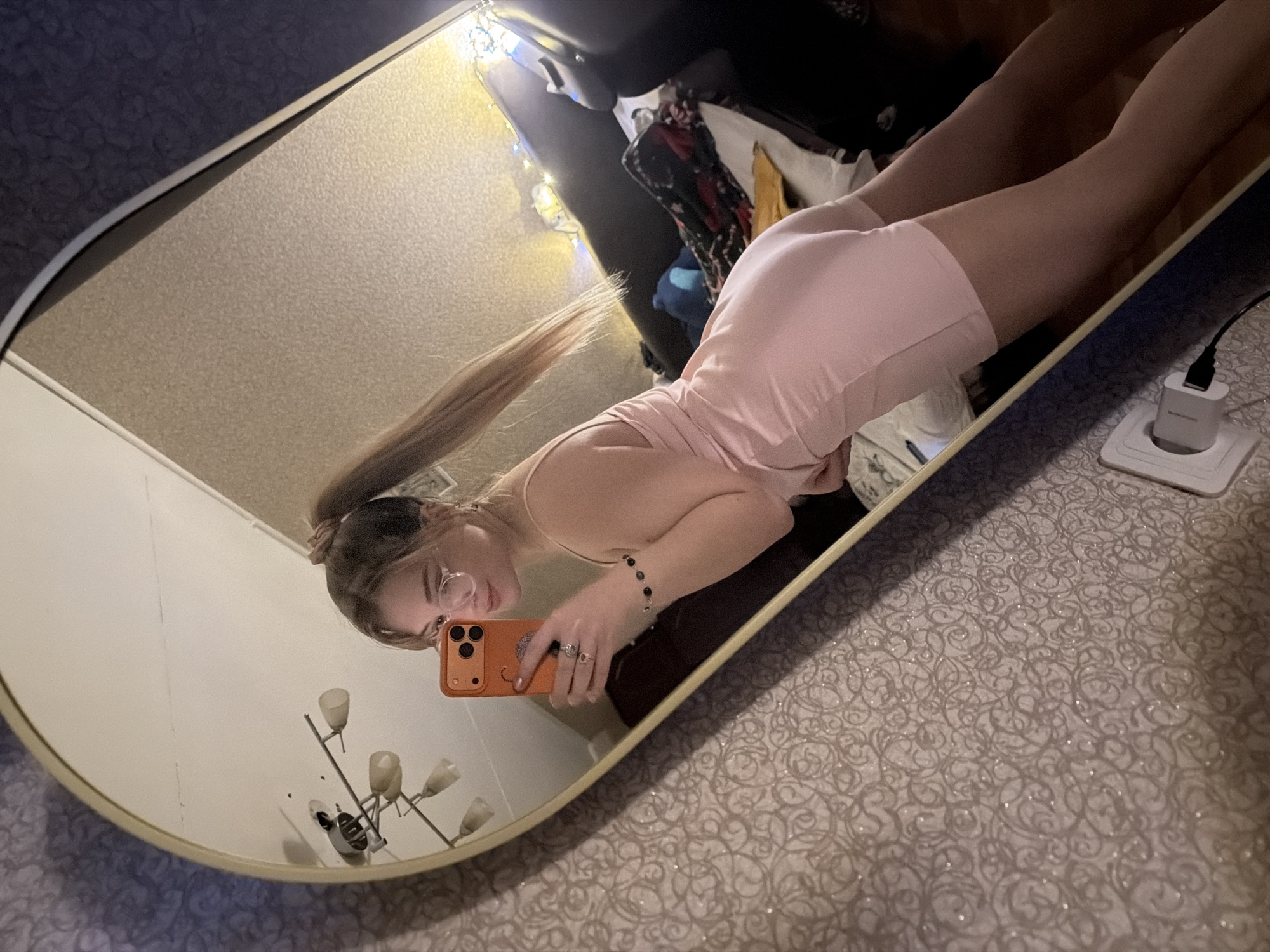 The image shows a woman taking a selfie in a mirror. The mirror has a gold frame and reflects a room interior. The woman is wearing glasses and a pink outfit. Her hair is in a high ponytail. She is holding an orange phone to take the picture. The reflection in the mirror also includes a floor lamp, clothes in a closet, and string lights. A charger is plugged into a wall outlet near the mirror. The bedding on which the mirror is placed has a patterned design. There is no information to discern the location in Naberezhnye Chelny, Russia.