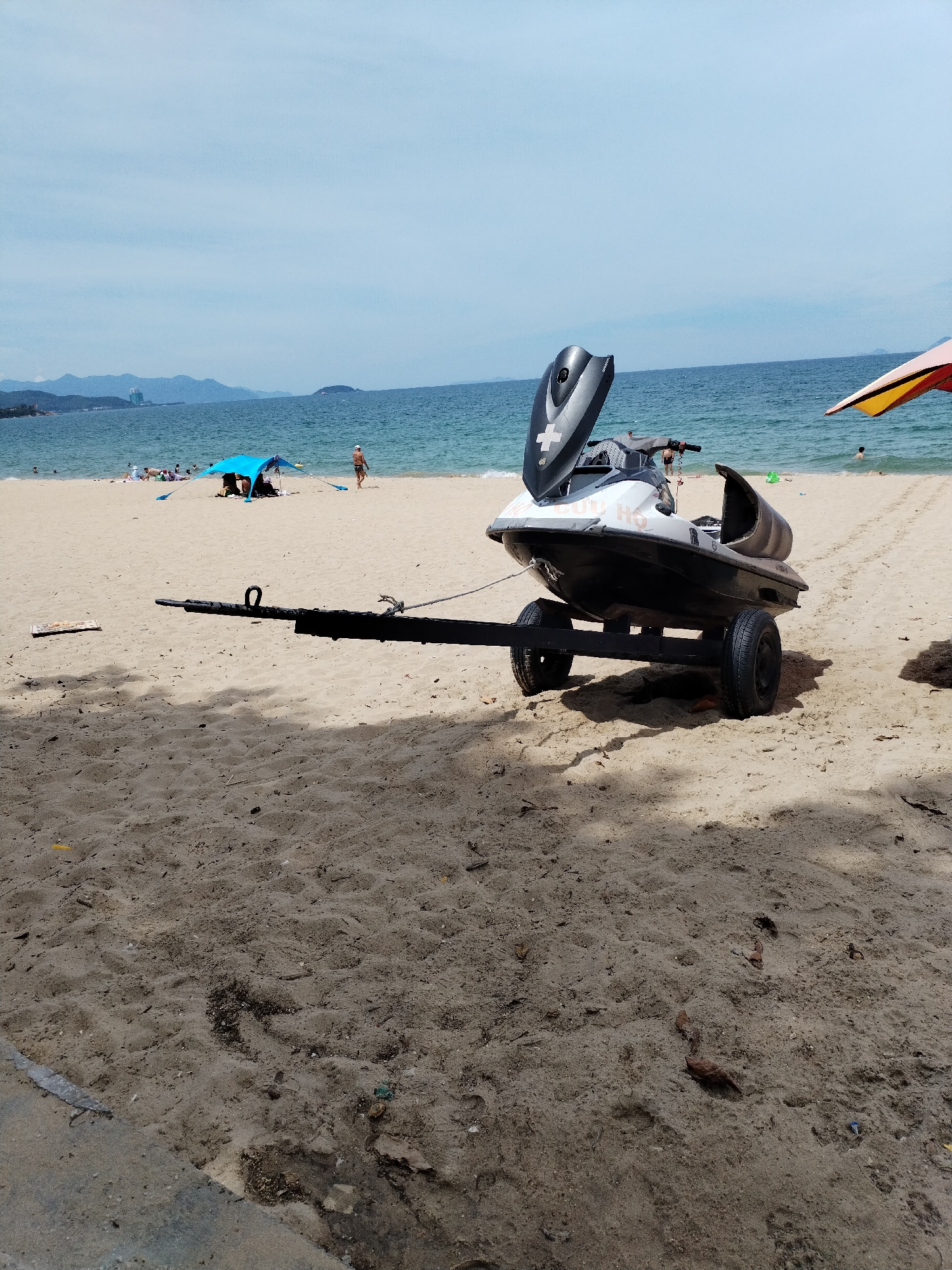A jet ski on a trailer is parked on a sandy beach in Nha Trang, Vietnam. The jet ski is black and white with a grey seat and has a white cross emblem on its front. The trailer has two black tires and a long black tow bar extending towards the left of the frame. In the background, the ocean is visible with a few people swimming and sunbathing on the beach. A blue tent is set up on the sand, and a colorful umbrella is partially visible on the right side of the image. The sky is clear and blue, with some distant mountains on the horizon. The overall scene depicts a typical beach day with recreational activities.