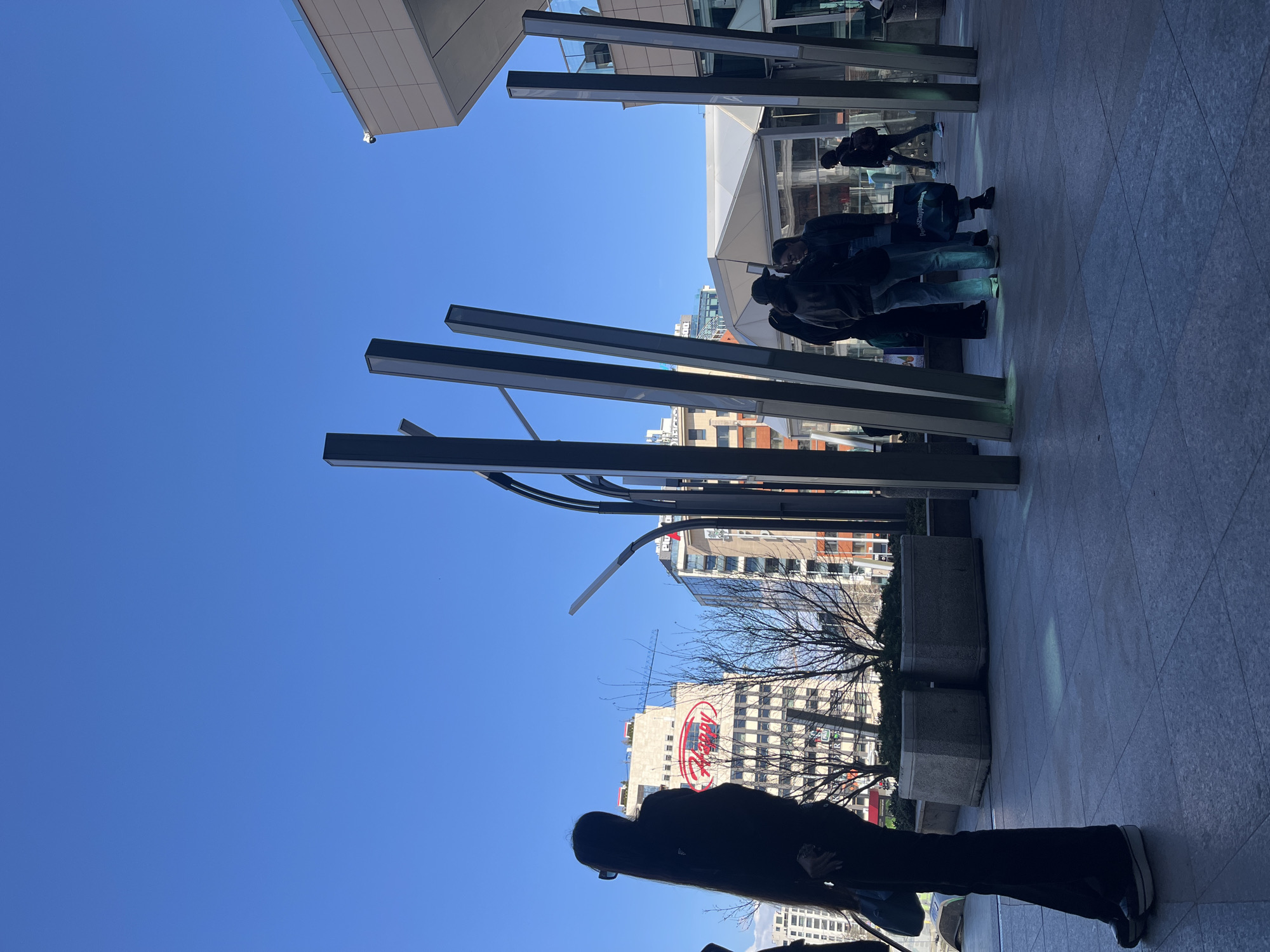 The image shows an outdoor scene in Sofia, Bulgaria. In the background, a tall building with a prominent "Happy" logo is visible. Foreground and mid-ground include structural elements like metal beams and what appear to be building facades. Several individuals are present; some are partially obscured, and others appear as silhouettes. The sky is clear and blue. The photograph's angle is unusual.