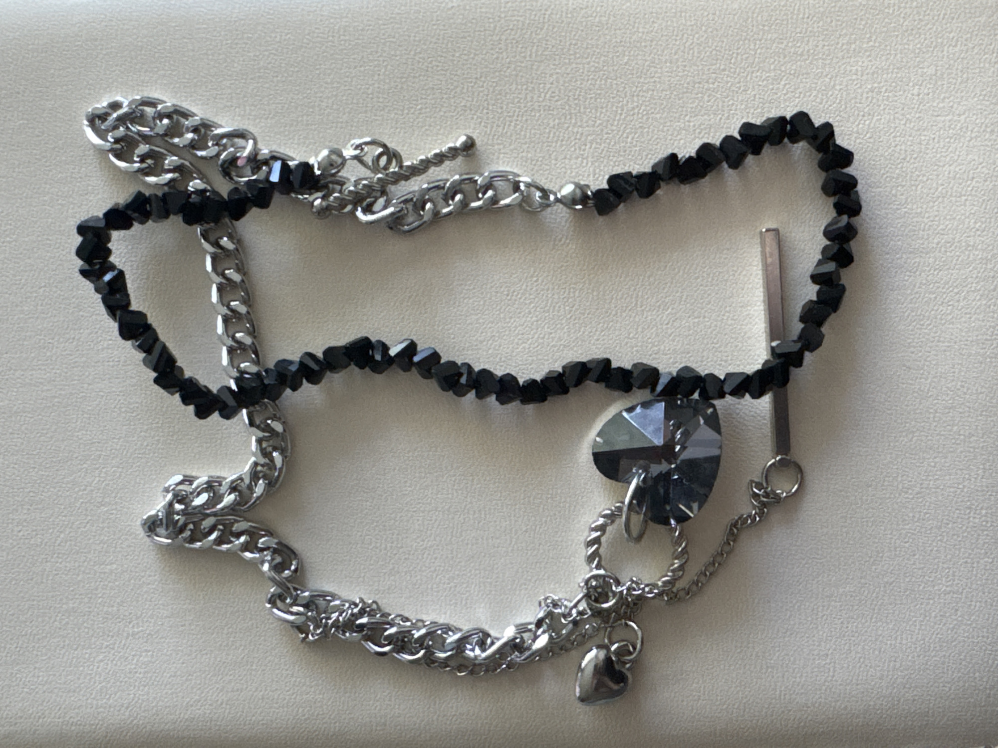 The image displays a close-up, overhead view of two necklaces arranged on a textured, light-colored surface, likely a tablecloth or a piece of fabric. The necklace on the left is a silver-toned chain with several charms: a small, smooth heart, a large faceted heart-shaped crystal pendant, and a smaller twisted loop with a jump ring. This necklace also features a delicate, finer chain. The necklace on the right is composed of small, faceted black beads strung together and a silver-toned bar charm. The necklaces are not located in the city of Яромінскі сельскі Савет, Belarus, as the image provides no geographical context.