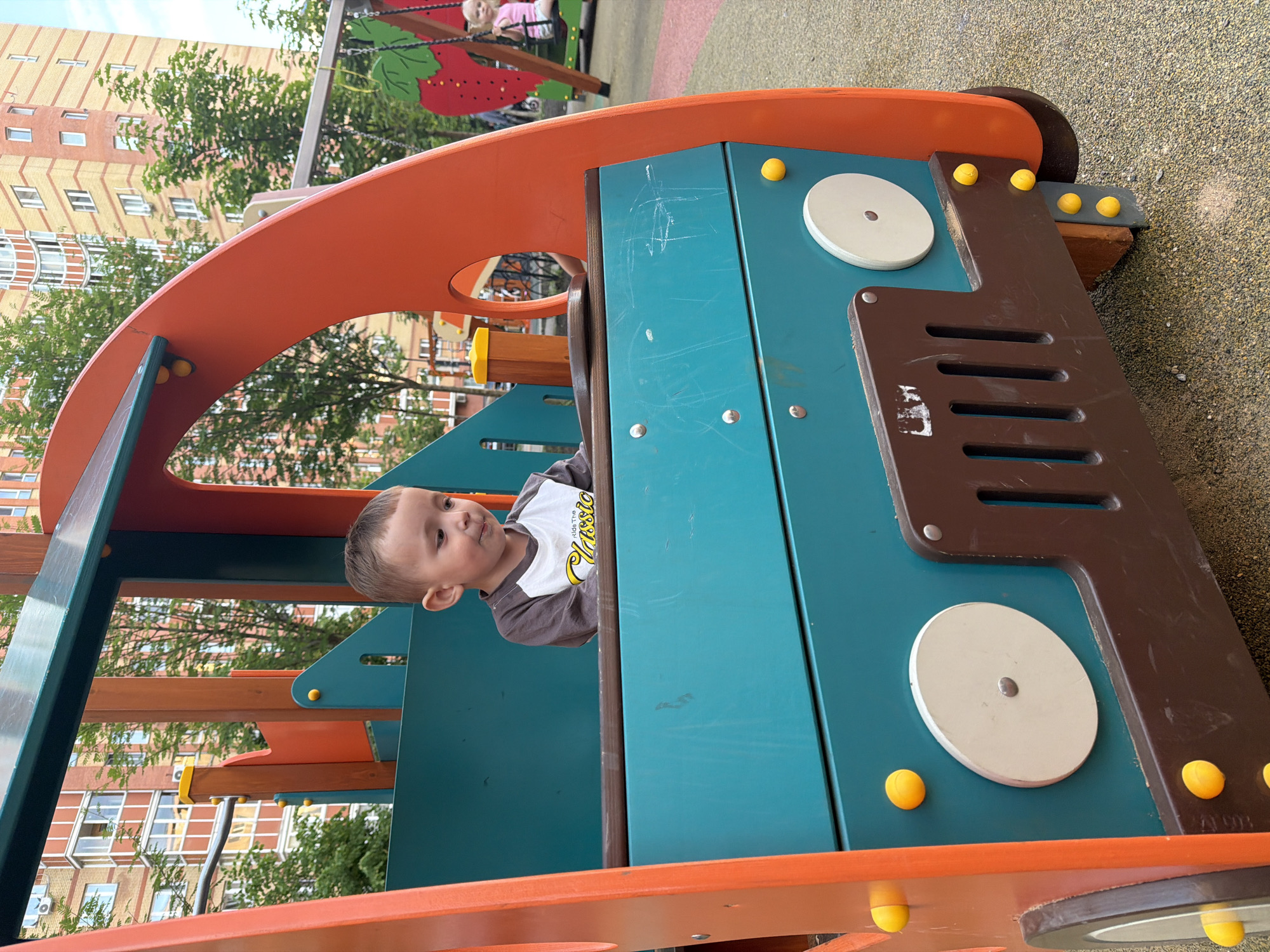 A young boy sits inside a colorful playground structure resembling a bus. The boy, with light brown hair and wearing a long-sleeved shirt with a graphic print, looks downwards. The playground structure is primarily teal and orange, with brown accents and yellow decorative elements. In the background, a multi-story building with red and yellow brickwork is visible, along with trees and a swing set with a child on it. The ground beneath the playground appears to be a soft, granular surface. The scene is outdoors on a cloudy day. This playground is located in the city of Vniissok, Russia.