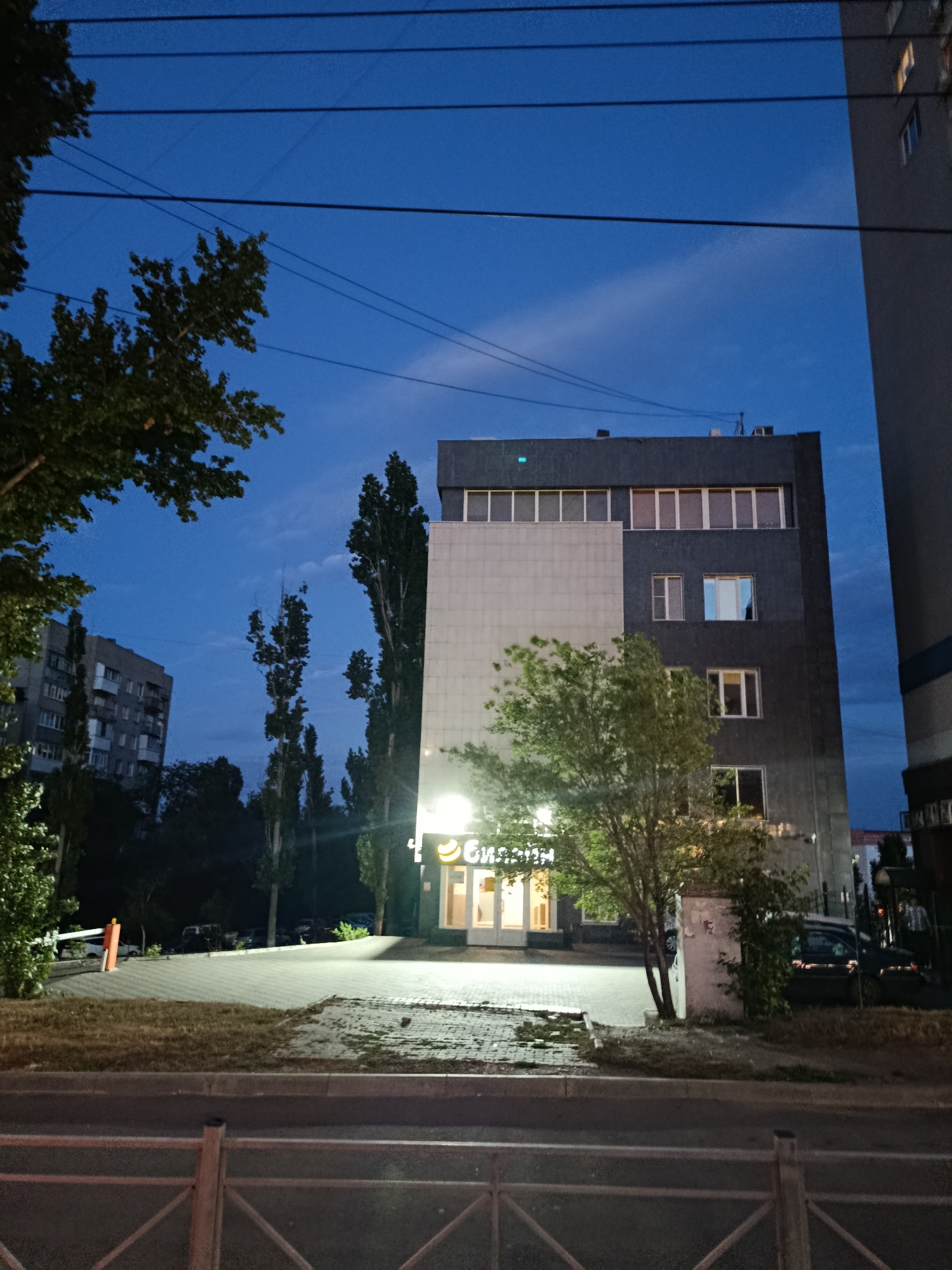 The image depicts a street scene at dusk in Saratov, Russia. In the foreground, a section of a metal fence runs horizontally across the bottom of the frame, with a road and a grassy strip in front of it. Behind the grass, a paved area with a slight incline is visible, with a white and orange barrier gate on the left.
The main subject is a multi-story building with a modern facade. The lower portion of the building features a light-colored, possibly stone or concrete, section with illuminated windows and a sign that reads "Биллиард" (Billiard) in white lettering on a yellow background. Above this is a darker section of the building with multiple windows, some of which are lit. To the right of this building, a sliver of another tall, gray building is visible, also with lit windows.
Several tall, slender trees stand in front of and around the buildings. The sky above is a deep blue, transitioning to lighter hues near the horizon, with some wispy clouds. In the background, another apartment building is visible to the left, and a few cars are parked in a lot behind the barrier gate. The overall atmosphere suggests an urban setting transitioning from day to night.
