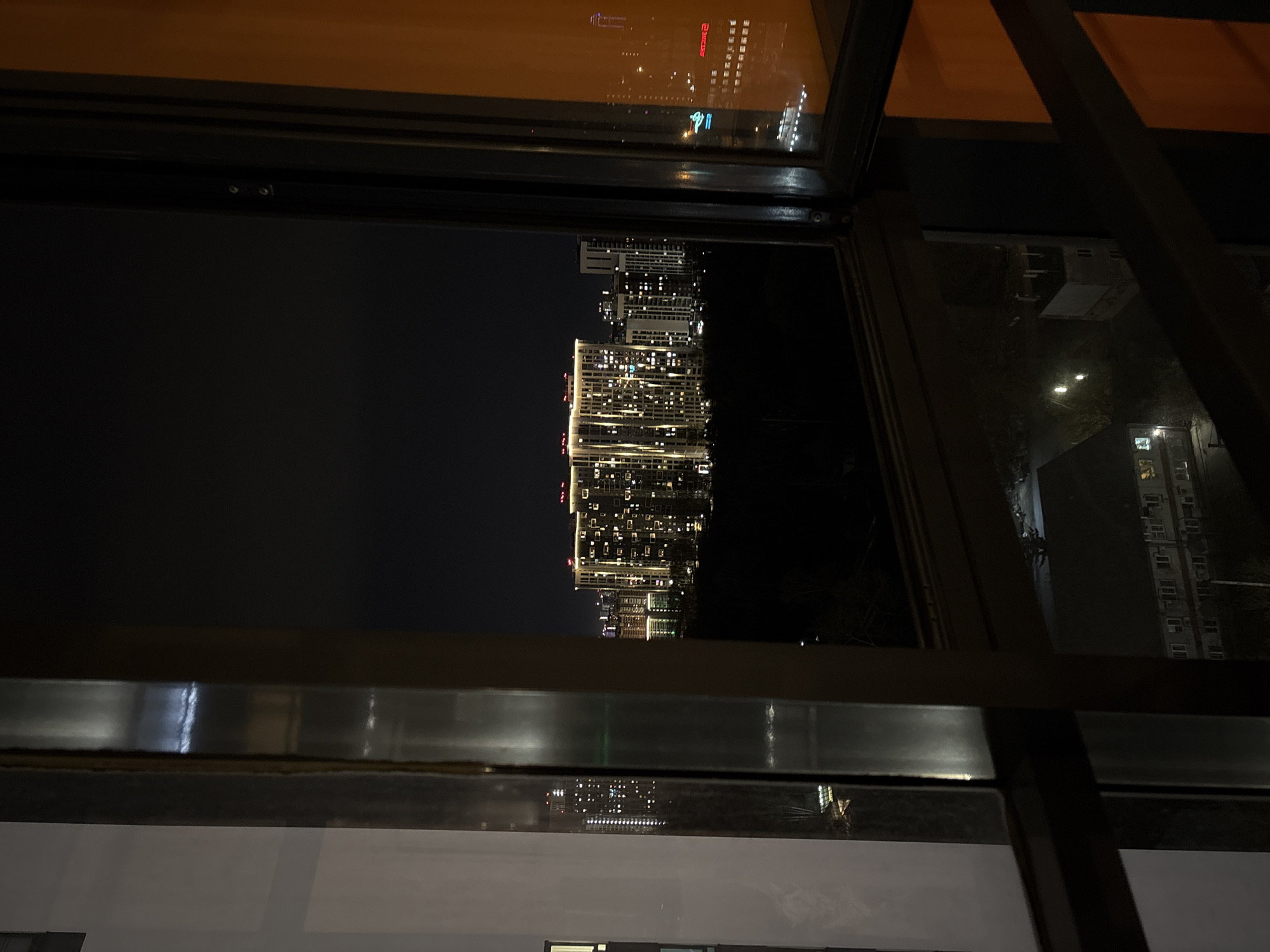 This media file captures a nighttime cityscape viewed from inside a building through a prominent window. The scene is dominated by a dense cluster of modern high-rise buildings, brightly illuminated by countless interior lights, suggesting a vibrant urban environment. The sky above the city is dark, confirming the late hour.
The view is framed by dark, thick window sashes, which cast strong reflections. In the upper portion of the window, a clear reflection shows an orange-brown interior ceiling and a reversed red sign featuring Cyrillic text. The visible words are "ДЕНЬГИ" (DENGY, meaning 'money') and "ЛЕТУЧИЕ" (LETUCHIYE, meaning 'flying' or 'volatile'). These Cyrillic characters serve as a significant location cue, pointing to a Russian-speaking area like Yekaterinburg, Russia. Further reflections are visible on the lower and side sections of the window, showing distorted elements of the city and possibly the interior of the room from which the photograph was taken.
No individuals are visible in the image. The focus is entirely on the architectural scale and the artificial glow of the city at night, creating a sense of urban expanse and modernity observed from a high vantage point.