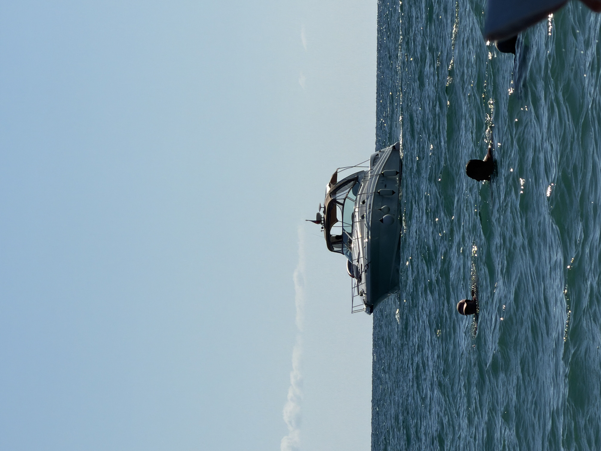 A wide shot captures a serene marine scene on a clear, sunny day, likely off the coast of Sochi, Russia. The sky is a pale, cloudless blue, meeting a shimmering turquoise sea that is moderately choppy. In the mid-ground, a sleek, white motor yacht with a canvas Bimini top cruises gracefully, leaving a gentle wake. Several inflatable fenders are visible along its hull. In the foreground, three individuals are swimming in the ocean. One person, wearing a dark baseball cap, is partially submerged, their head and shoulders visible above the water. To their right, another person is also swimming, their head visible. A third person, with only their head visible, is further to the right and closer to the edge of the frame. The sunlight glints off the water's surface, creating sparkling highlights. The image evokes a sense of leisure and outdoor recreation.