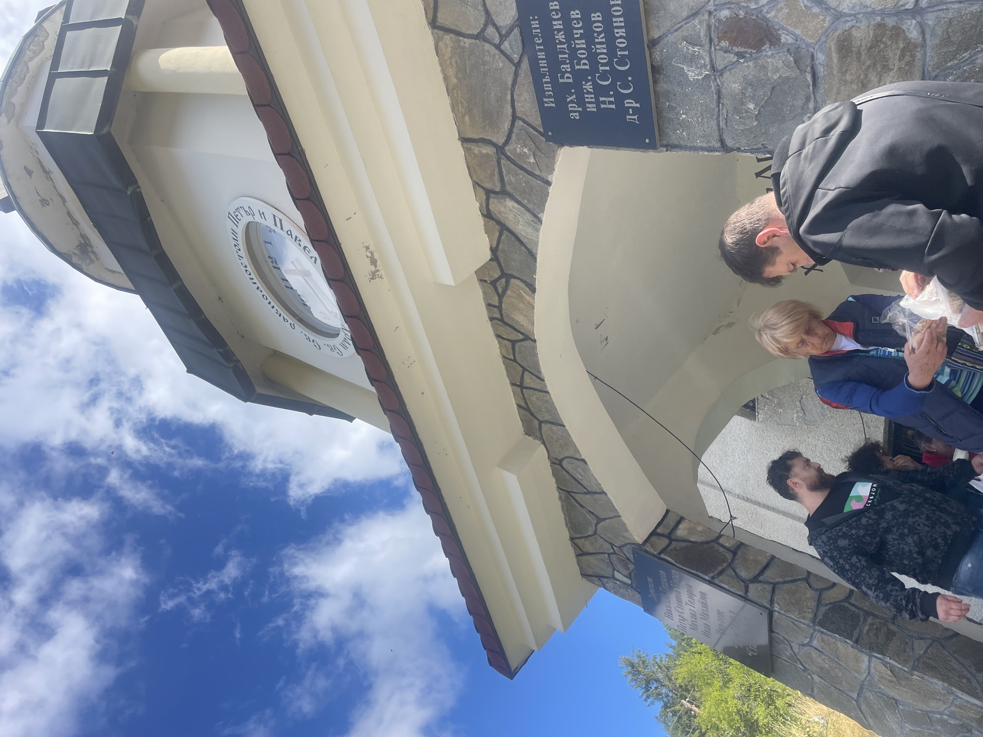 The image depicts an outdoor scene in front of a stone and white stucco building with a clock tower. The sky is blue with scattered white clouds. In the foreground, three people are visible. A man with a beard wearing a patterned sweatshirt and jeans is walking towards the left. To his right, a woman with blonde hair wearing a blue jacket is holding a plastic bag. Beside her, a person in a black hoodie is also holding a plastic bag.
On the building, there are two plaques. The left plaque, mounted on stone, has text in Bulgarian. The right plaque, also in Bulgarian, is mounted on stone above an archway and lists names and titles, including "Изпълнители:" (Executors:), "арх. Балджиев" (arch. Baldzhiev), "инж. Бойчев" (eng. Boychev), "Н. Стойков" (N. Stoykov), and "д-р С. Стоянов" (Dr. S. Stoyanov). The clock face on the tower has Roman numerals and writing in Cyrillic.
The building appears to be a religious structure, possibly a church or monastery, located in the city of Manastir, Bulgaria, given the presence of Bulgarian text and context. The overall atmosphere suggests a public gathering or event.