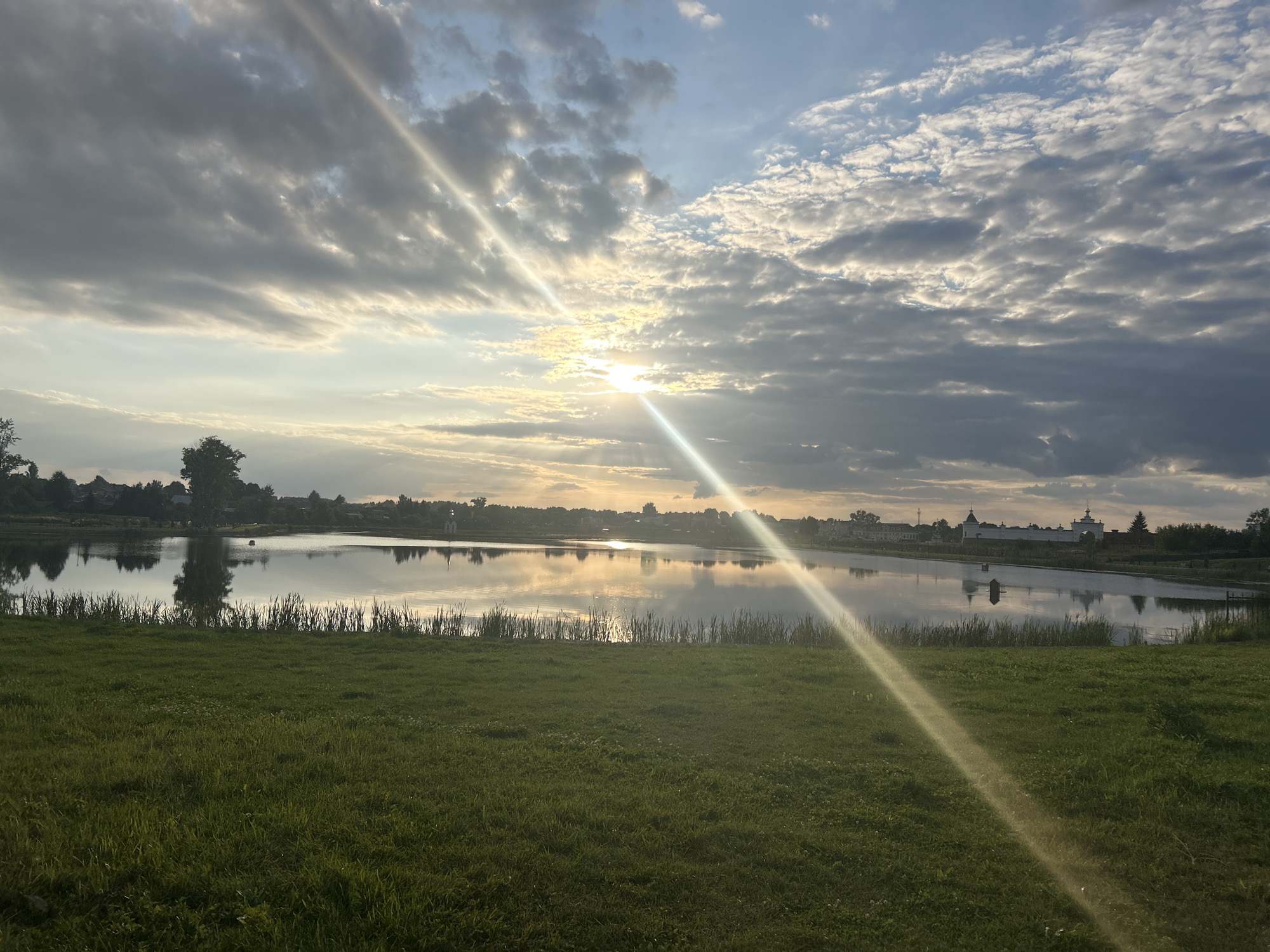 The image depicts a serene landscape scene with a body of water reflecting the sky. In the foreground, a grassy field stretches out, meeting the edge of the water. A line of reeds or tall grass borders the water. Across the water, a distant shoreline is visible, featuring a cluster of buildings with distinct architectural elements, including domes that suggest a religious or historical site. Trees are scattered along the shoreline. The sky is partially cloudy, with sunlight breaking through, creating bright rays that stream across the scene and reflect on the water's surface. The overall impression is one of tranquility and natural beauty, possibly at sunrise or sunset, with the distant structures implying a settlement or monastic complex, consistent with the known landmarks in Diveyevo, Russia.