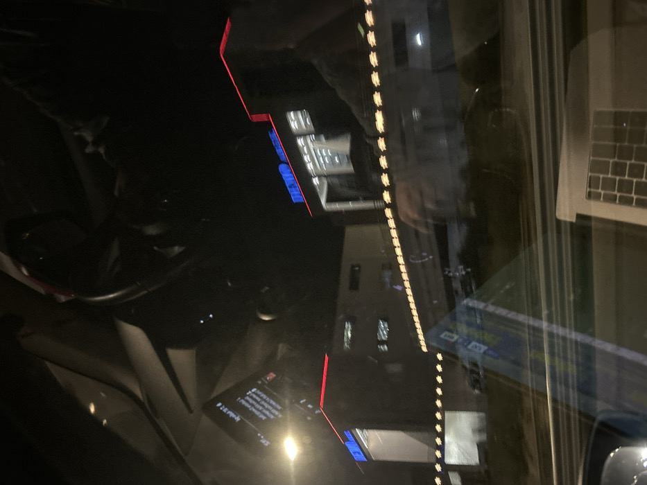 The media file captures a rotated (90 degrees clockwise) nighttime scene viewed from inside a vehicle, dominated by reflections on the glass. The dark environment is illuminated by various artificial lights.
Inside the vehicle, a portion of a steering wheel is visible in the bottom-left corner. Faint reflections of a person's hands and upper body are discernible, particularly on the right side near a reflected laptop keyboard and screen. A reflected display, likely a smartphone or car screen, shows text in the bottom-center.
Through the glass, several buildings are visible in the background, featuring illuminated windows with white and blue lights. A prominent vertical line of evenly spaced orange-yellow lights stretches across the right side of the image. Additionally, red linear lights appear at the top and bottom edges, possibly outlining architectural features or signage.
The activity depicts a static moment, likely from a parked or stopped vehicle at night. The visible text on the reflected screen, partially upside down, includes "S-mobile", "1. Σύνδεση" (1. Login), "2. Εγγραφή" (2. Register), and "3. Ξεχάσατε τον κωδικό;" (3. Forgot password?). These Greek phrases, combined with the overall setting, place the scene in Agios Stefanos, Greece.
