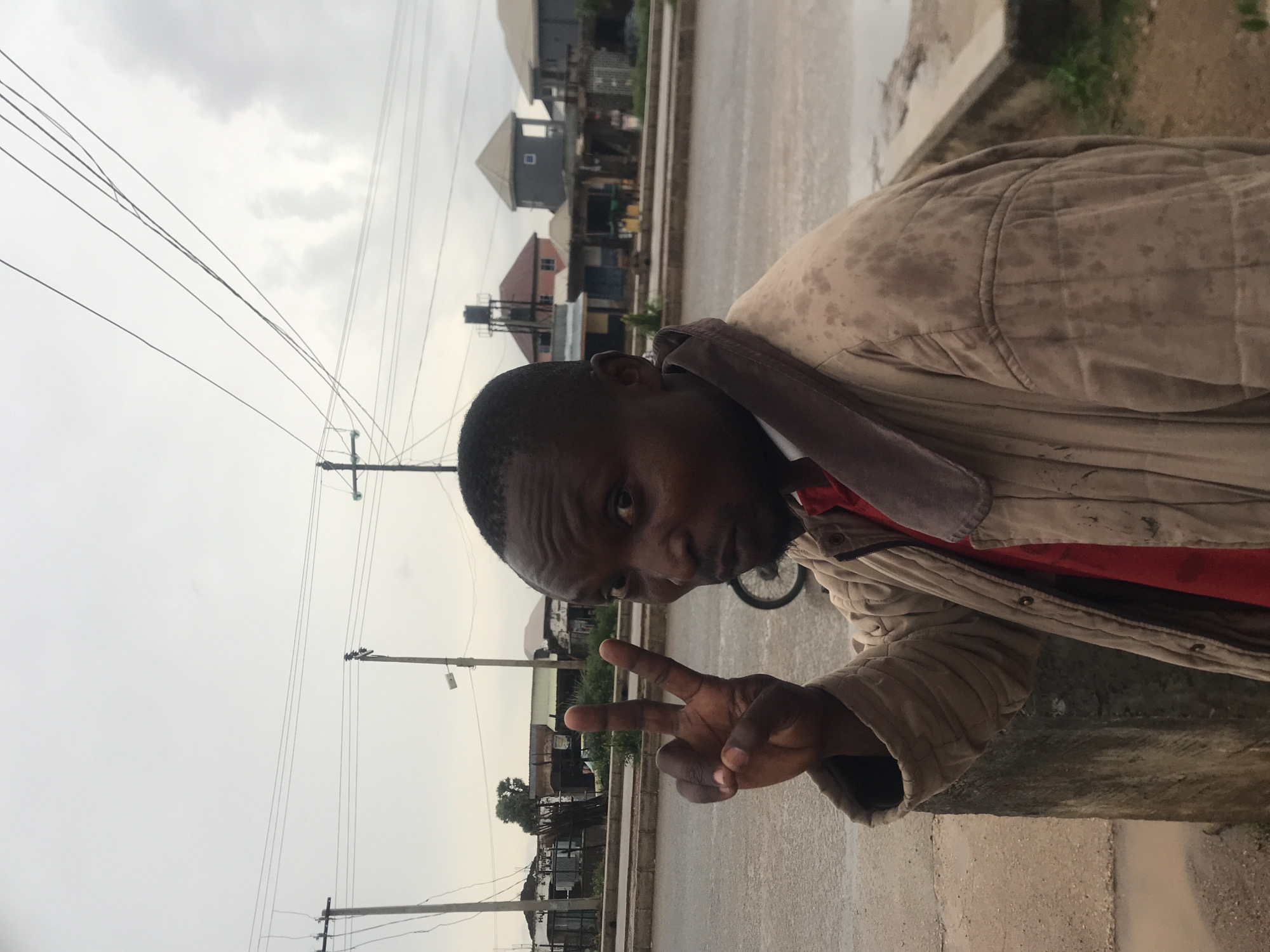 The image, taken in Rafin Par, Nigeria, captures a man posing outdoors in an urban or semi-urban setting. The photo is oriented vertically, showing the horizon running up the right side of the frame.
The primary subject is a man, positioned prominently in the foreground. He looks directly at the camera with a neutral to slightly curious expression. He is wearing a light-colored, possibly tan or beige, quilted jacket over a red shirt, with the jacket appearing somewhat soiled or stained. His right hand is raised, forming a "peace" or "victory" sign with two fingers extended. He has short hair and some facial stubble.
The background reveals an overcast sky, suggesting a cloudy day. Numerous utility poles and a complex network of power lines span across the upper and middle sections of the frame. Below the lines, a road or street is visible, appearing damp, potentially from recent rain. A concrete curb runs along the edge of the road. In the distance, several buildings of varying sizes and architectural styles are scattered, indicating a residential or mixed-use area. A bicycle wheel is partially visible behind the man on the right side of the image. The overall atmosphere suggests a casual moment captured in a local street setting under cloudy weather conditions. No visible text is present in the image.