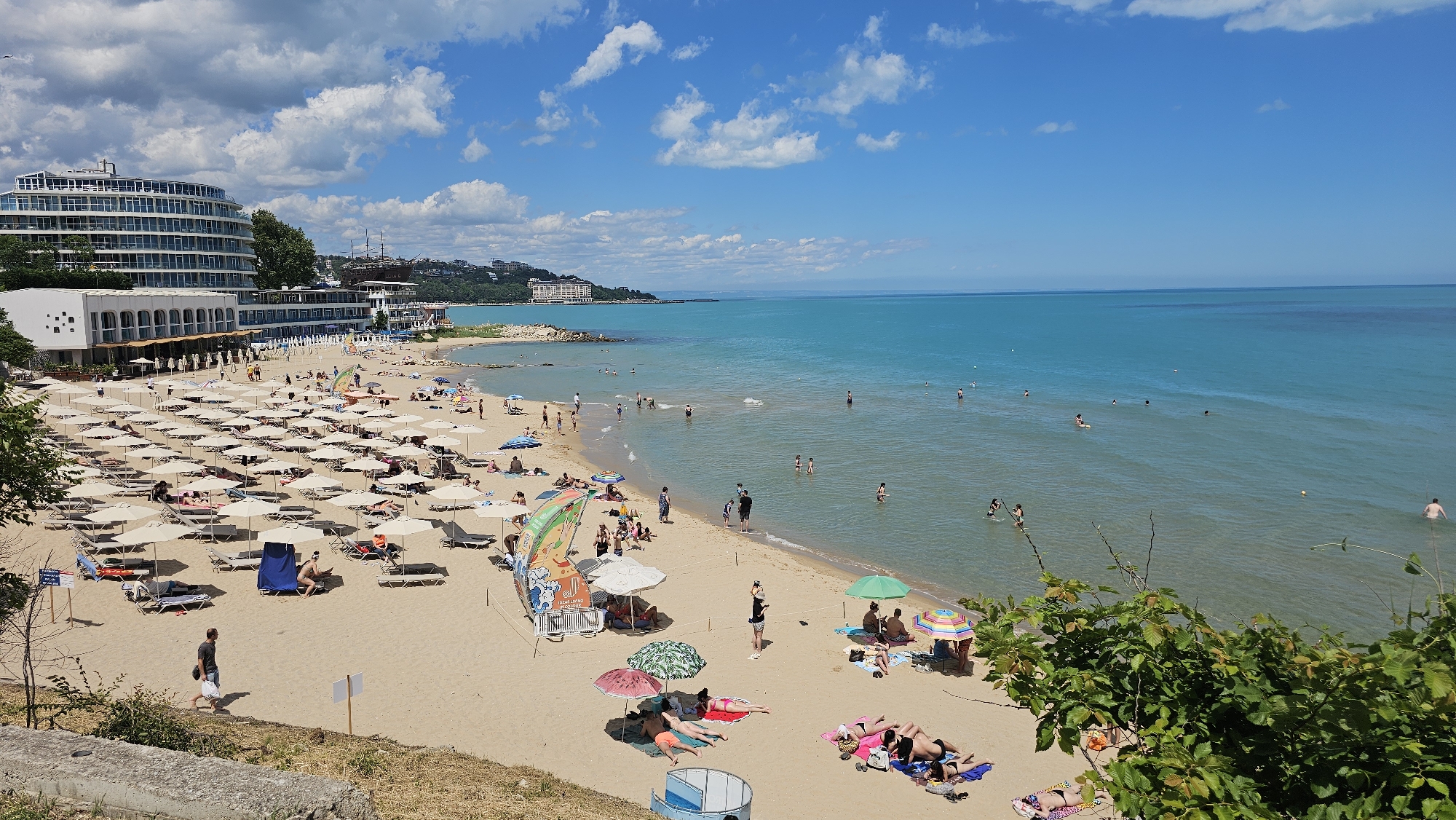 This is an aerial photograph of the Sunny Beach in Varna, Bulgaria, on a clear day with a few clouds in the sky. The beach is filled with many people sunbathing and swimming in the bright blue sea. Numerous beach umbrellas and lounge chairs are set up on the sand, indicating a popular tourist destination. A large, modern hotel building with a curved facade is visible on the left side of the image, overlooking the beach. In the distance, more buildings and the coastline can be seen. The sea is calm and inviting, with several individuals enjoying the water. The scene captures a vibrant and lively summer atmosphere on the Bulgarian coast.