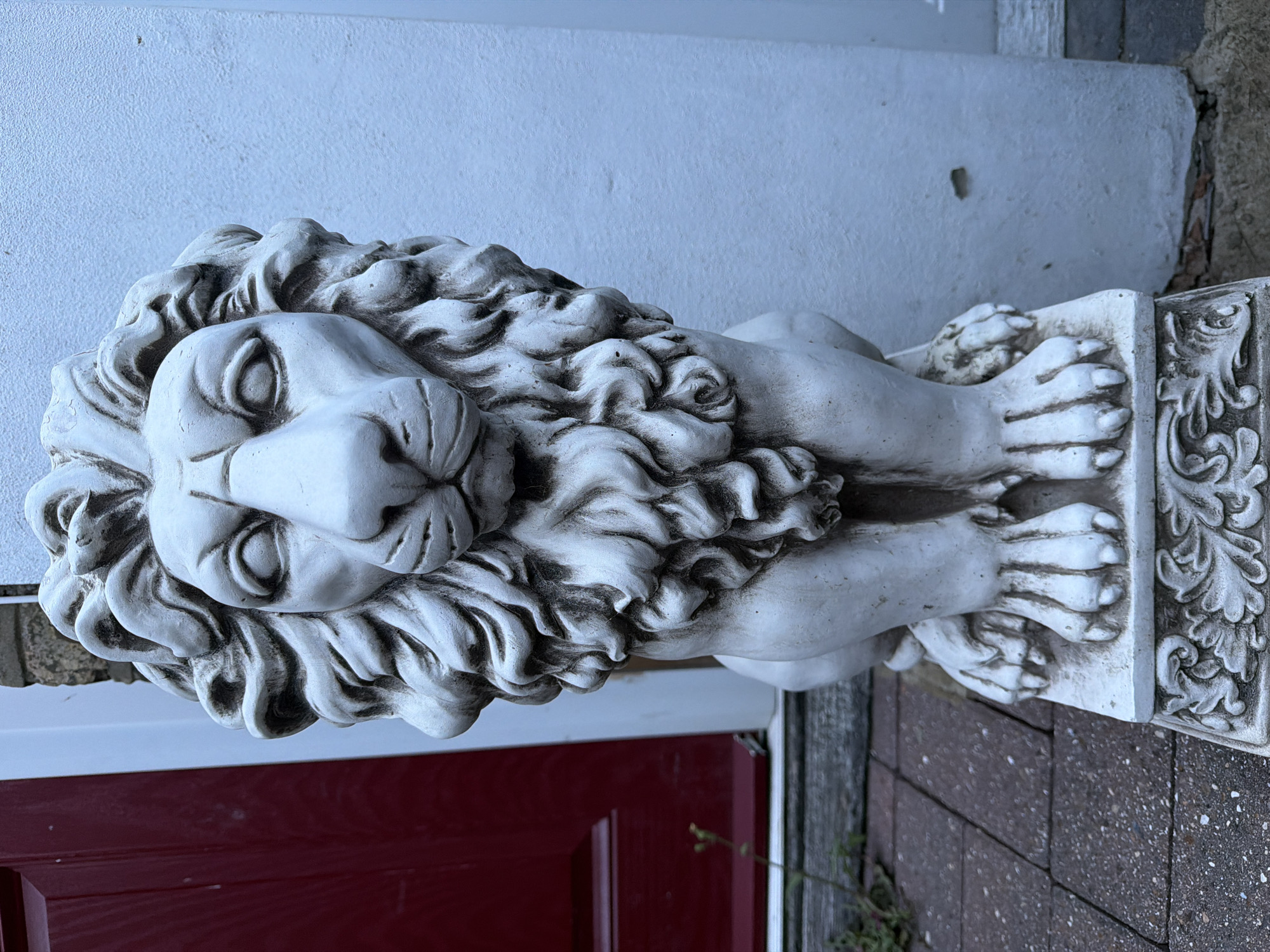 The scene is situated in Maidstone, United Kingdom, and depicts a weathered white lion statue positioned outdoors, adjacent to a building entrance. The statue, crafted from a light-colored material such as concrete or cast stone, is seated upright and faces directly forward. It features a highly detailed, curly mane encompassing its head and shoulders, pronounced facial attributes including closed eyes, a distinct nose, and a discernible mouth. Its front paws, equipped with visible claws, rest upon a rectangular base adorned with an ornate, possibly acanthus leaf, relief pattern.
To the left, a section of a deep red, paneled door is visible, indicating a portal to a structure. Below the door, a dark-colored threshold or step is present, with minor green plant growth visible along its base and in the foreground. To the right of the statue, a light-colored, textured wall, likely stucco or painted masonry, extends vertically, displaying minimal surface blemishes. The ground in the immediate vicinity of the statue is composed of reddish-brown paving stones with a rough surface texture. The lighting is diffused and uniform, suggesting an overcast ambient condition. The image is devoid of any human presence or discernible action.
