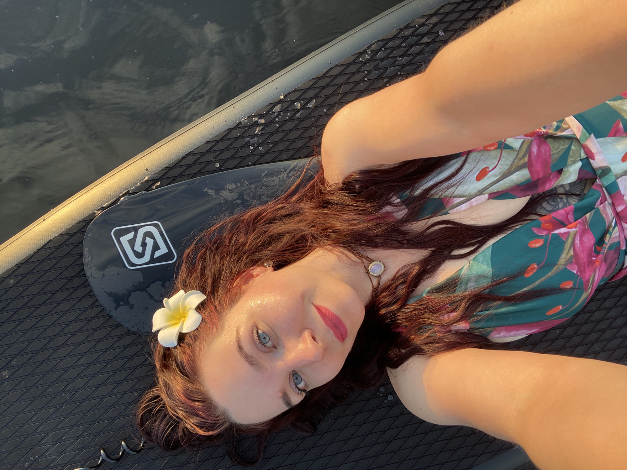 A woman lies on a black paddleboard, possibly on a body of water near Chaikovsky, Russia. She has red hair and blue eyes, wearing a floral dress and a flower in her hair. Her arms are raised as if taking a selfie. The paddleboard has a logo on it and water droplets are visible. The background shows a dark, reflective surface, presumably water.