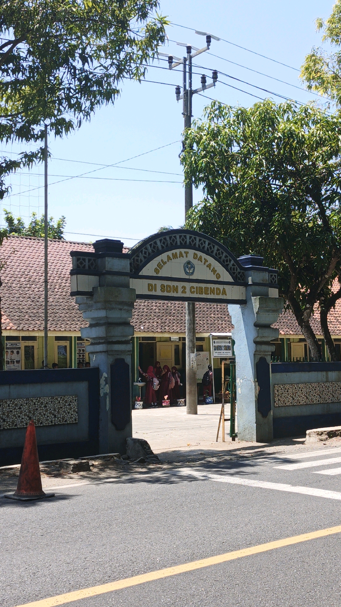The image shows the entrance to an Indonesian elementary school, identified by the sign above the archway reading "SELAMAT DATANG DI SDN 2 CIBENDA". The school building has a red tiled roof and light yellow walls. Several students, dressed in maroon uniforms, are gathered near the entrance, some with backpacks. A traffic cone is visible on the left side of the road, in front of the school. The scene appears to be in a semi-urban or rural area given the presence of trees, power lines, and a paved road with a pedestrian crossing. The sky is clear and blue, indicating a sunny day.