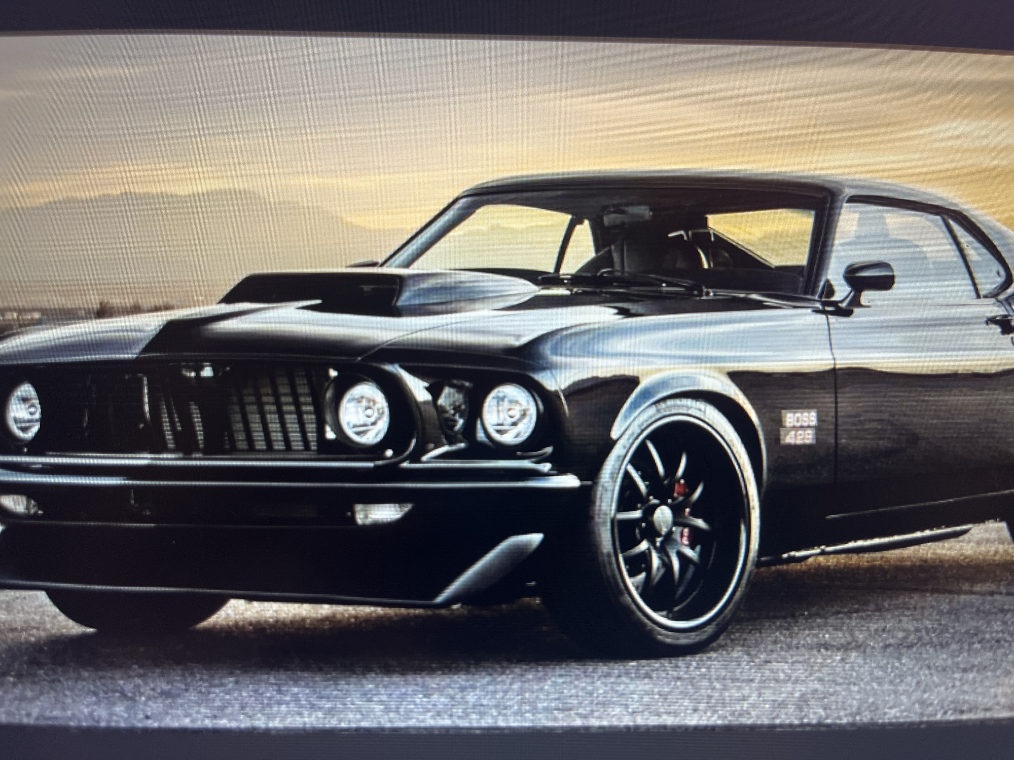 A high-resolution, slightly pixelated photograph displays a classic black Ford Mustang Boss 429, angled to reveal its front and driver's side. The car dominates the lower two-thirds of the frame, showcasing its aggressive styling. Notable features include a prominent raised hood scoop, a dark horizontal bar grille, and dual circular headlights on each side, with the inner pair appearing smaller, possibly as fog lights. The car is fitted with large, multi-spoke black custom wheels, through which red brake calipers are visible. Twin chrome side exhaust pipes extend along the lower body, from behind the front wheel well. A "BOSS 429" emblem is clearly visible on the front fender.
The setting is outdoors, with the car positioned on a dark, textured paved surface, likely a road or an overlook. In the background, distant, layered mountains stretch across the horizon, illuminated by a warm, golden light that suggests either a sunrise or sunset. The sky above is a gradient of warm, soft hues transitioning from golden-orange near the mountains to a lighter, hazy grey-blue higher up. The atmosphere appears clear and calm. There are no people visible, allowing the powerful aesthetic of the car and the serene natural landscape to be the sole focus. The overall mood is one of appreciation for classic automotive design amidst a scenic backdrop.