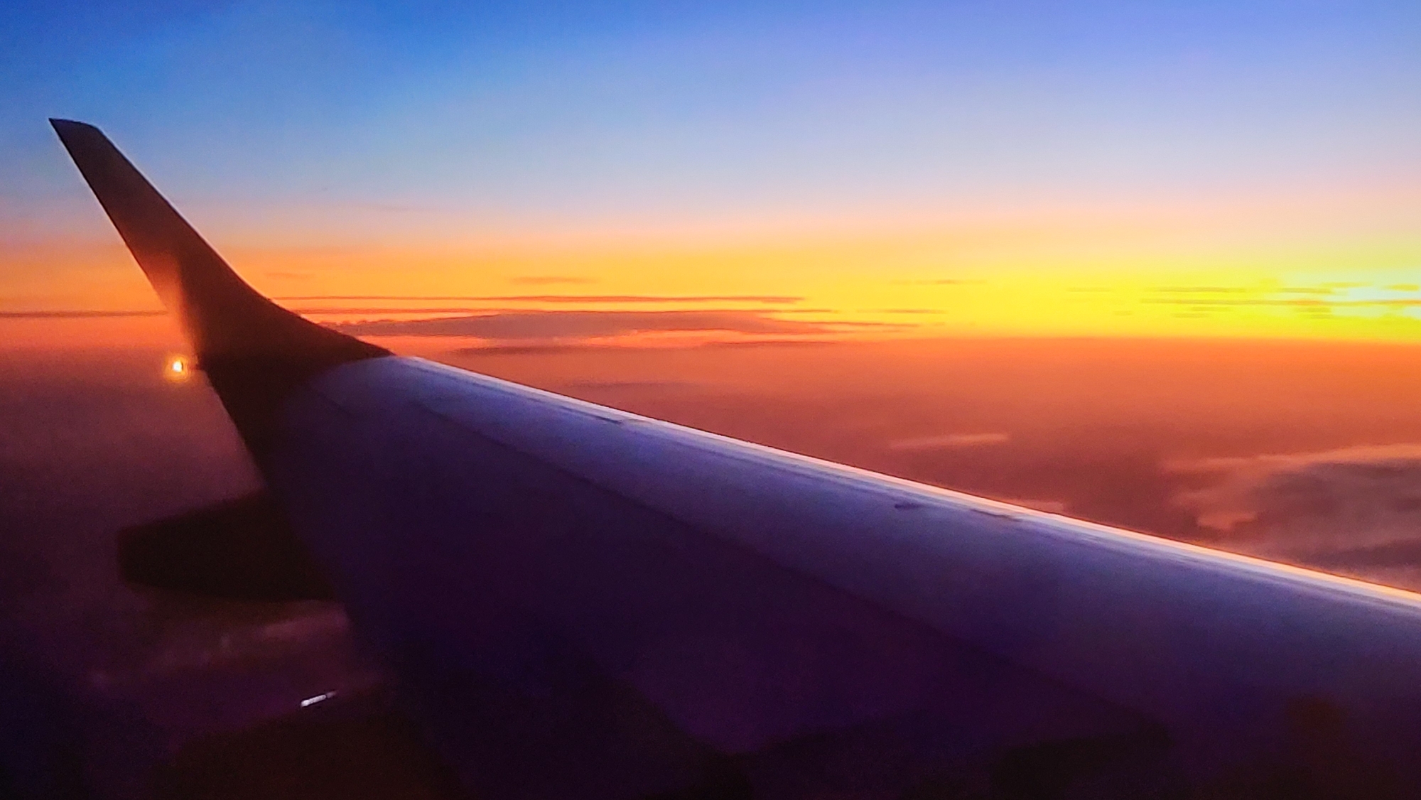 The image depicts an airplane wing viewed from inside the aircraft during sunset. The wing is in the foreground, partially obscuring the view of the horizon. The sky transitions from blue at the top to yellow and orange near the horizon. There is a band of clouds visible on the horizon. There are no discernible people, subjects, actions, or interactions present in the image. The location in Yerevan, Armenia, cannot be determined from the image.