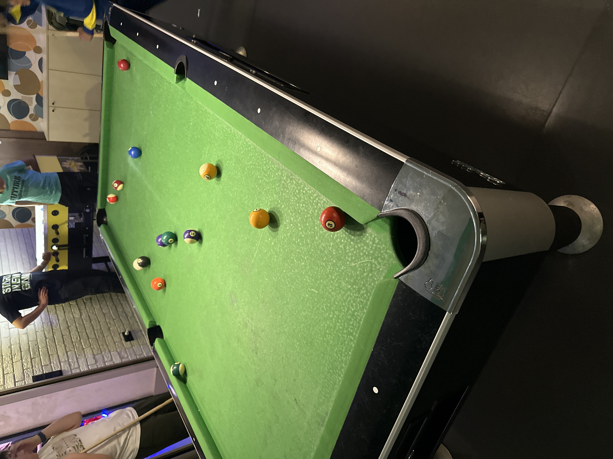 A rotated, high-angle shot captures an active game of billiards (pool) within an indoor setting, likely an entertainment venue. The central focus is a green-felted pool table, scattered with numerous pool balls.
On the table, several balls are visible: a solid red 8-ball positioned near a corner pocket, a solid blue ball, and a solid yellow ball. Striped balls include a yellow and white, a red and white, a purple and white 12-ball, a green and white 10-ball, a burgundy and white 15-ball, and an orange and white 13-ball. The felt shows signs of use, with visible chalk residue or wear. A prominent chrome-colored corner pocket is in the foreground.
Several people are engaged with the game. In the bottom left, a person wearing a white t-shirt holds a pool cue, appearing to take a shot. To their right, another individual in a black t-shirt is partially visible, with the text "START NEW GAME" clearly readable. A third person, wearing a light blue t-shirt with "FUTURE" printed on the front, stands near the table on the left side. A fourth person, partially obscured in the top left, wears a blue and yellow jacket.
The background features a wall adorned with a modern circular pattern on the upper left, transitioning to a light-colored, brick-textured wall below. A light-colored cabinet is also visible. The artificial lighting suggests an indoor evening or night setting. The scene depicts a social gathering centered around the game of pool.