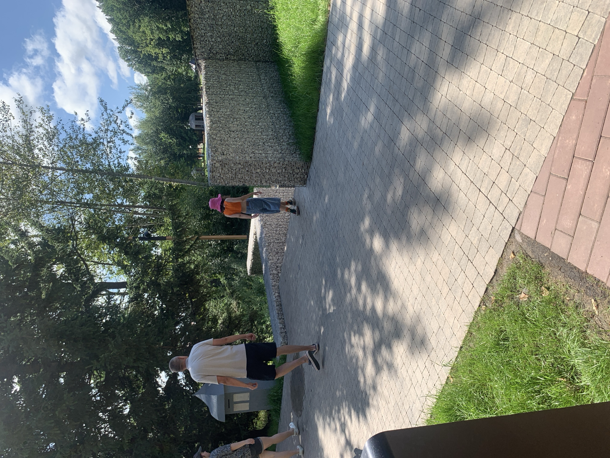 The image shows a paved walkway in a park-like setting. On the left, a man in a light-colored t-shirt and dark shorts walks away from the camera. Beside him, another person, partially visible, is also walking away. Further down the path, a young person wearing an orange crop top, denim shorts, and a pink bucket hat is walking away from the camera. The walkway is made of light-colored interlocking pavers, with a section of red brick pavers in the foreground. To the right of the walkway is a gabion wall filled with stones, with green grass beyond it. In the background, there are trees and a blue sky with white clouds. A dark trash receptacle is visible in the lower left corner. There is no information in the image to indicate the location is Arbuzov-Baran, Russia.
