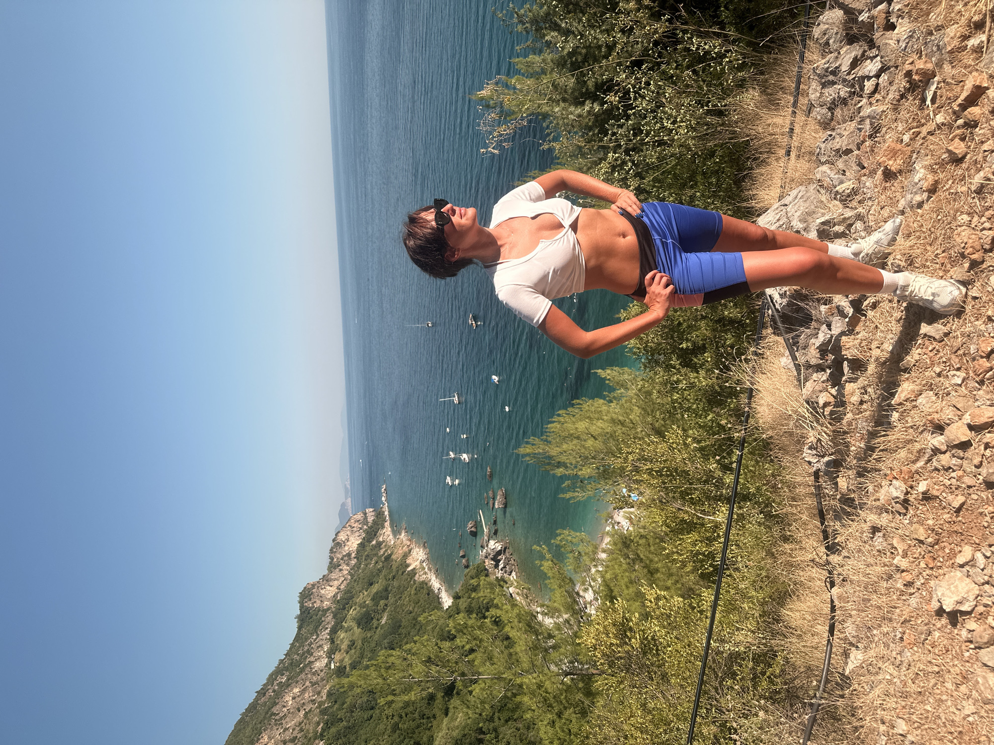 A woman stands on a rocky, overgrown hillside overlooking a turquoise sea. She is wearing a white cropped top and blue athletic shorts with black and pink accents. White sneakers and white socks complete her outfit. She has short, dark hair and is wearing sunglasses. Her hands are on her hips, and she is looking out at the sea. In the distance, sailboats dot the water, and a rugged, green coastline stretches into the distance. The sky is a clear, bright blue. The location is Prijevor, Montenegro.